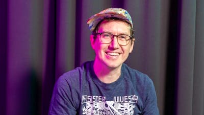 A smiling man wearing glasses and a colorful cap, seated in front of a dark curtain.