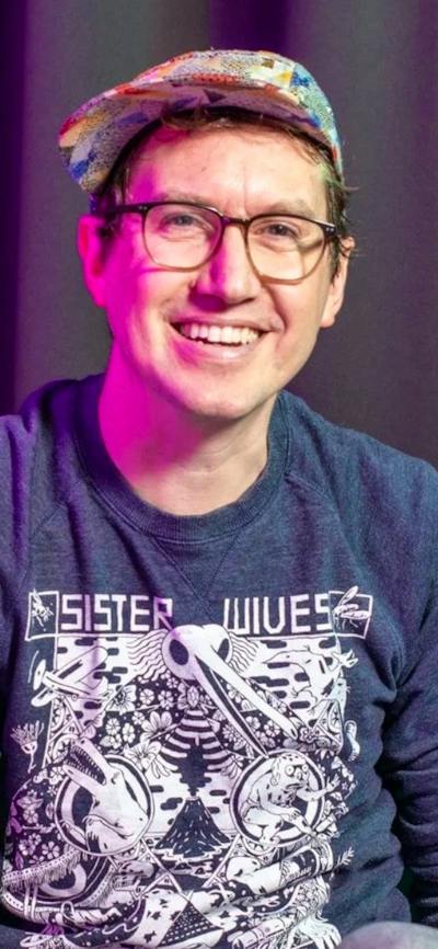 A smiling man wearing glasses and a colorful cap, seated in front of a dark curtain.