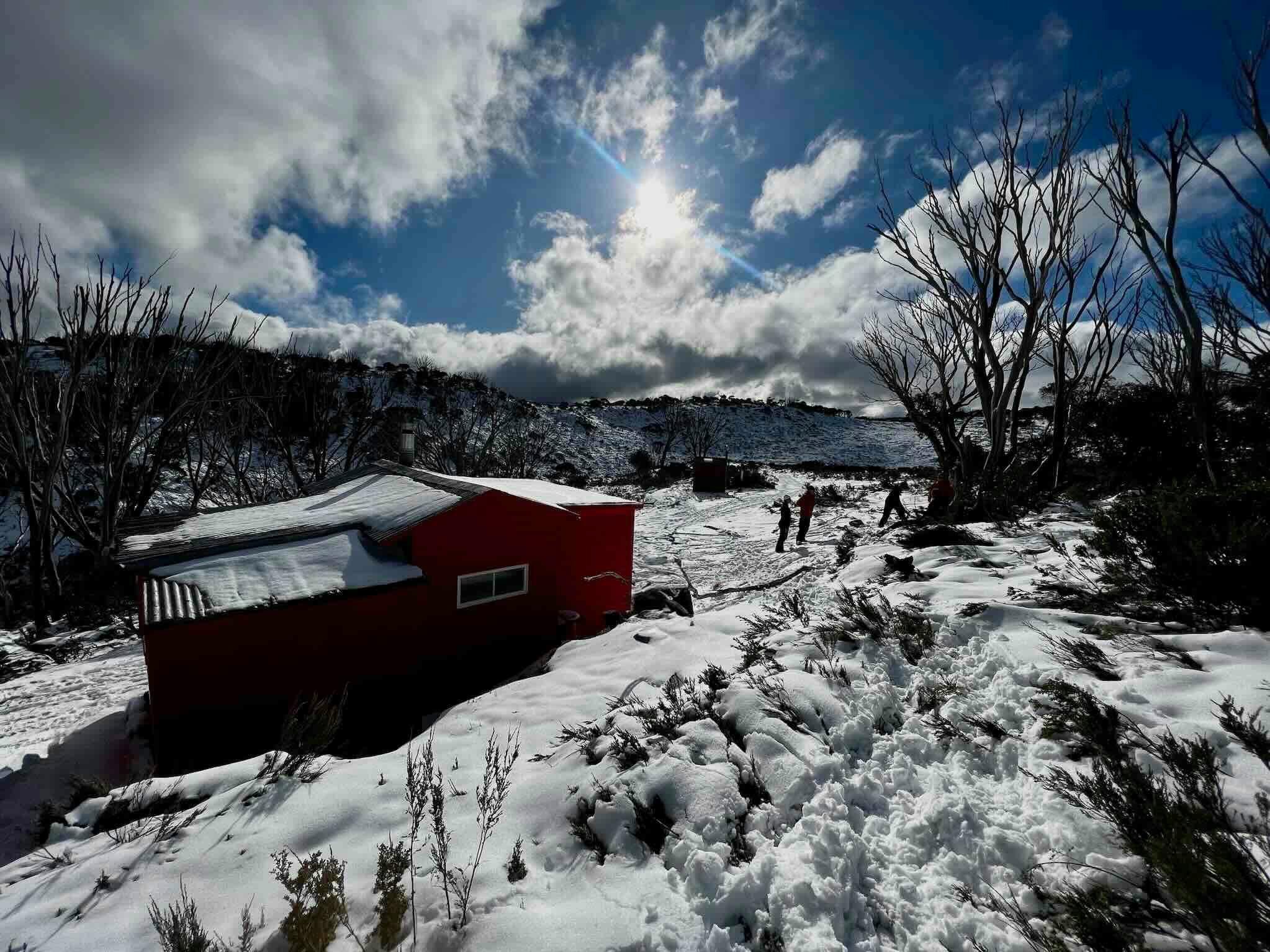 A red hut in the mountains covered in snow with hikers in the backcountry and the sun shining.