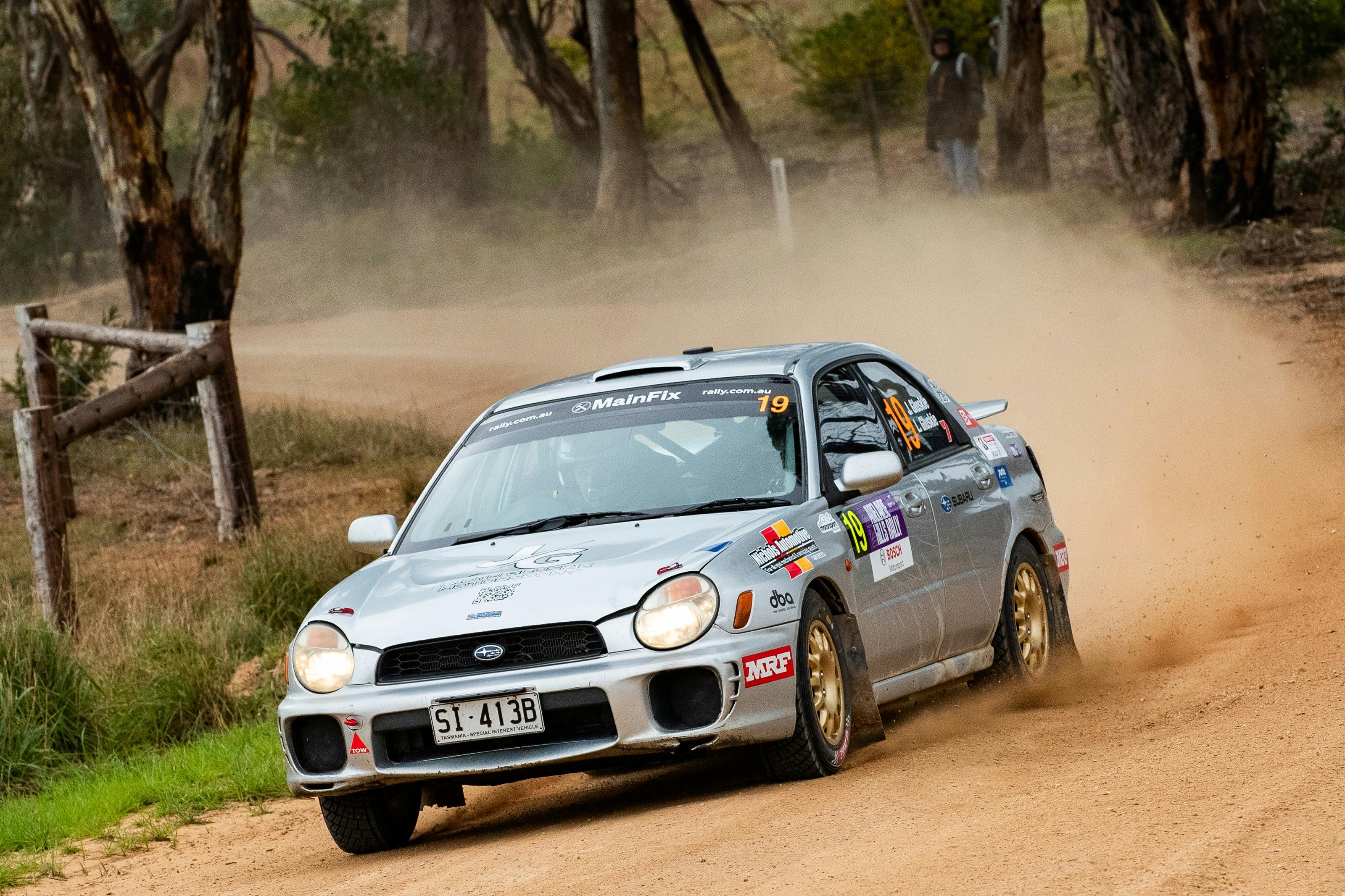 Silver rally car is driving on gravel road with dust kicked up behind it.