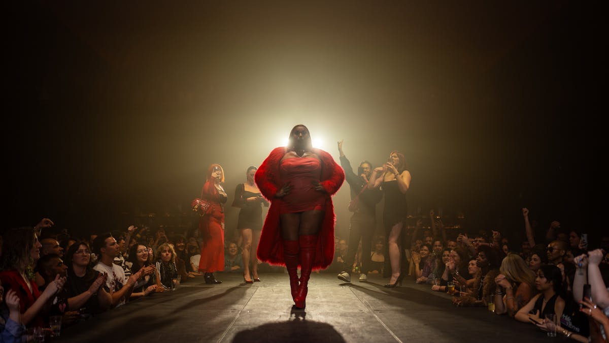 A performer in a red dress is backlit as they walk down a runway