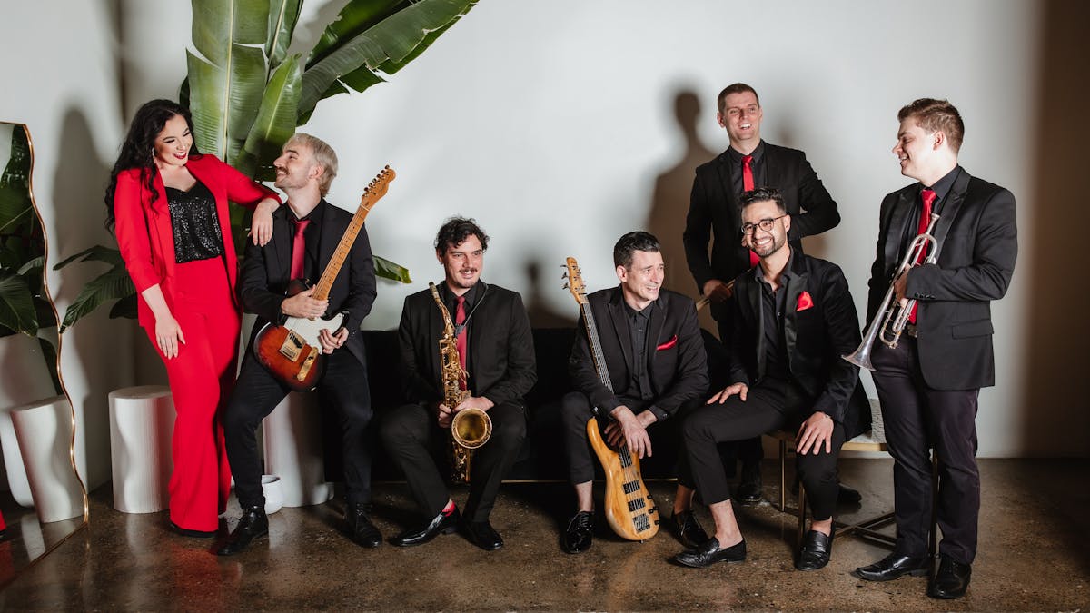6 musicians are arranged on and around a couch. They laugh with each other. The mood is light,