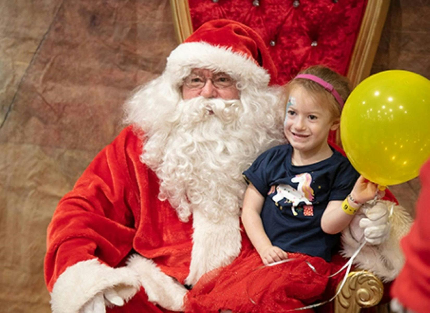 Santa sitting in his chair with a young girl on his lap holding a yellow balloon