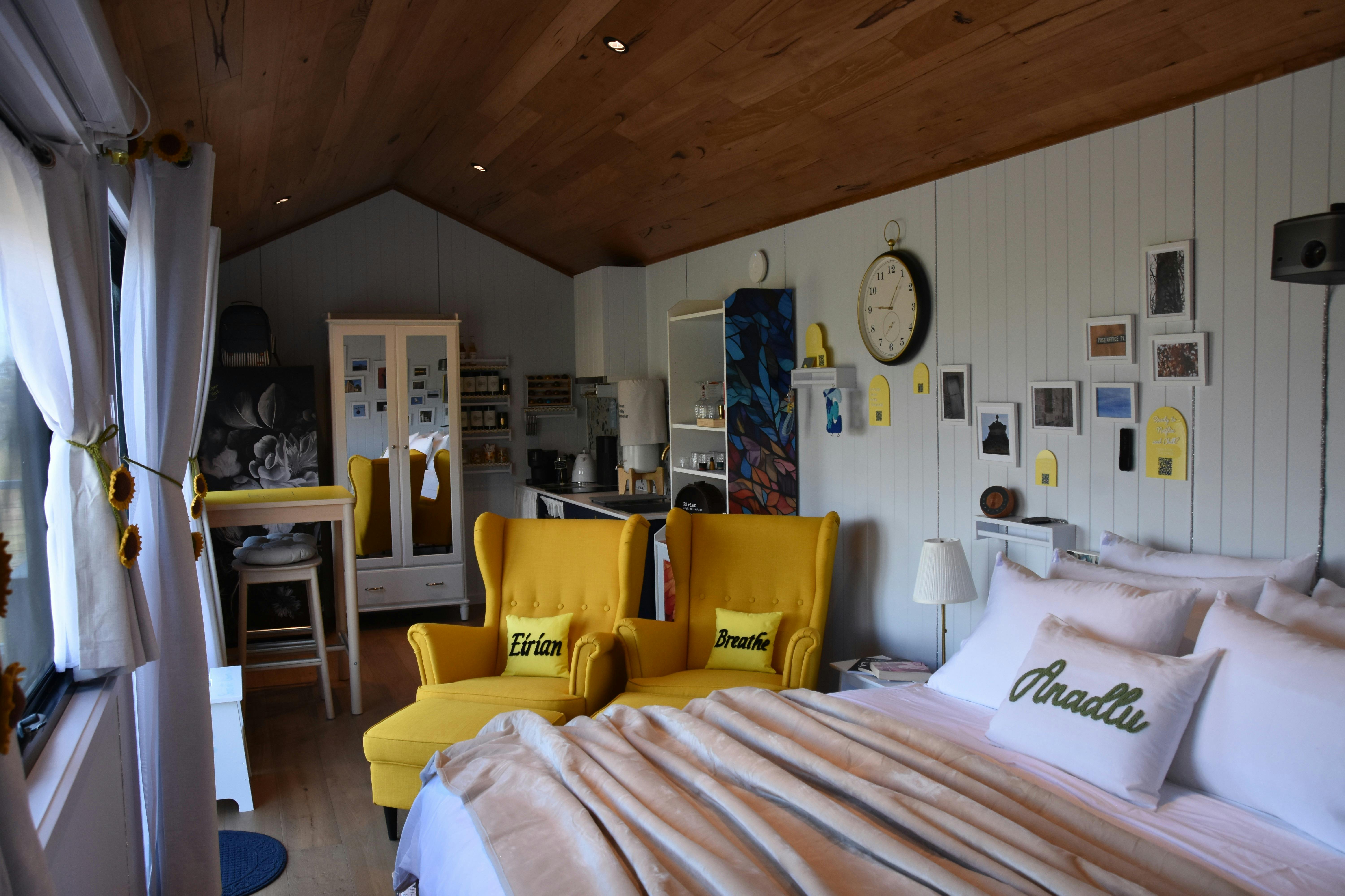 Interior of tiny home with bed, yellow chairs, and timber ceiling.