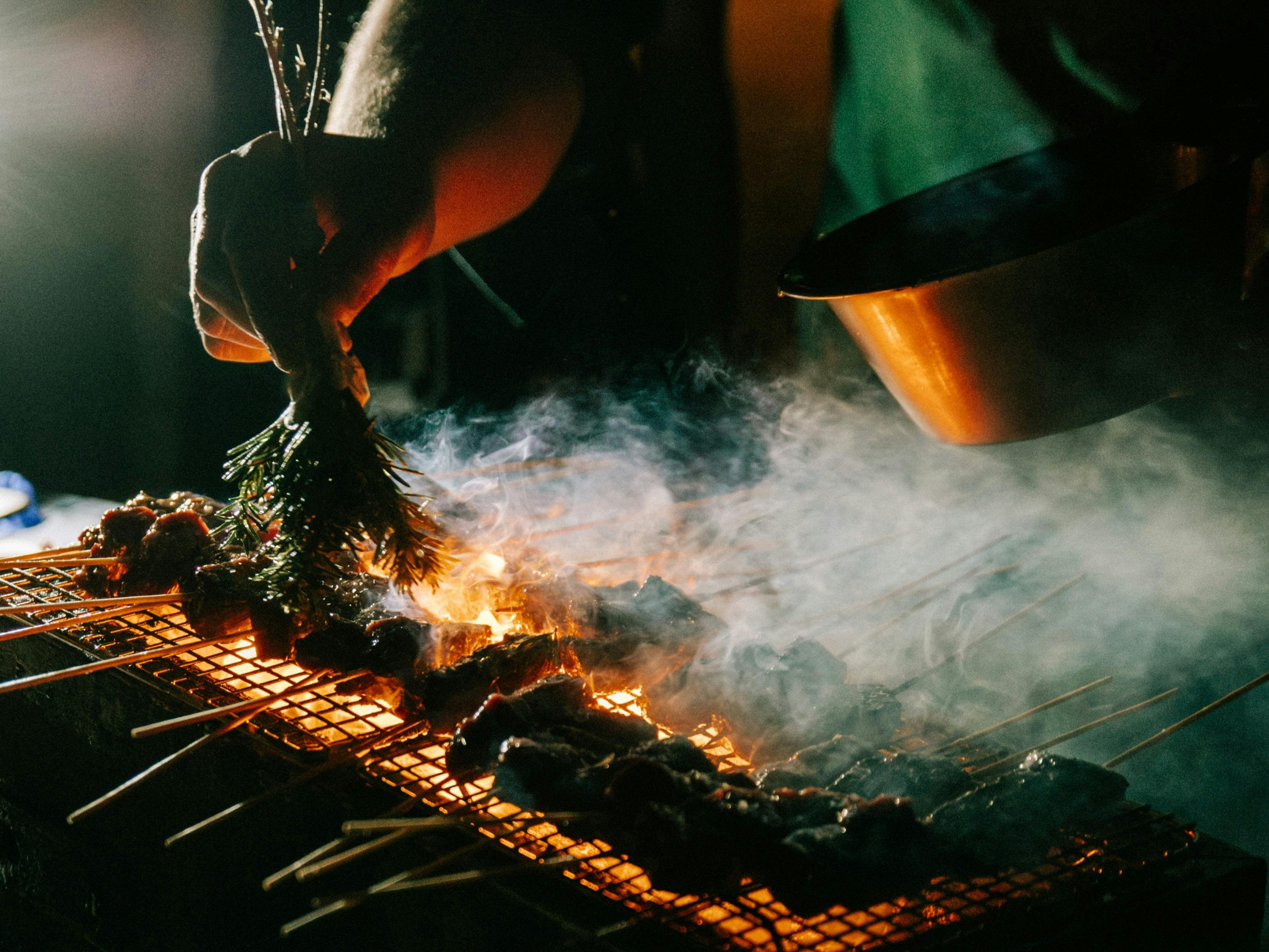 A chef brushes herbs on meat on a lit bbq, low lighting