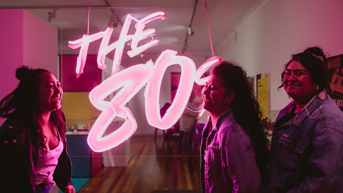 Three women standing beside pink illuminated sign 'The 80s'