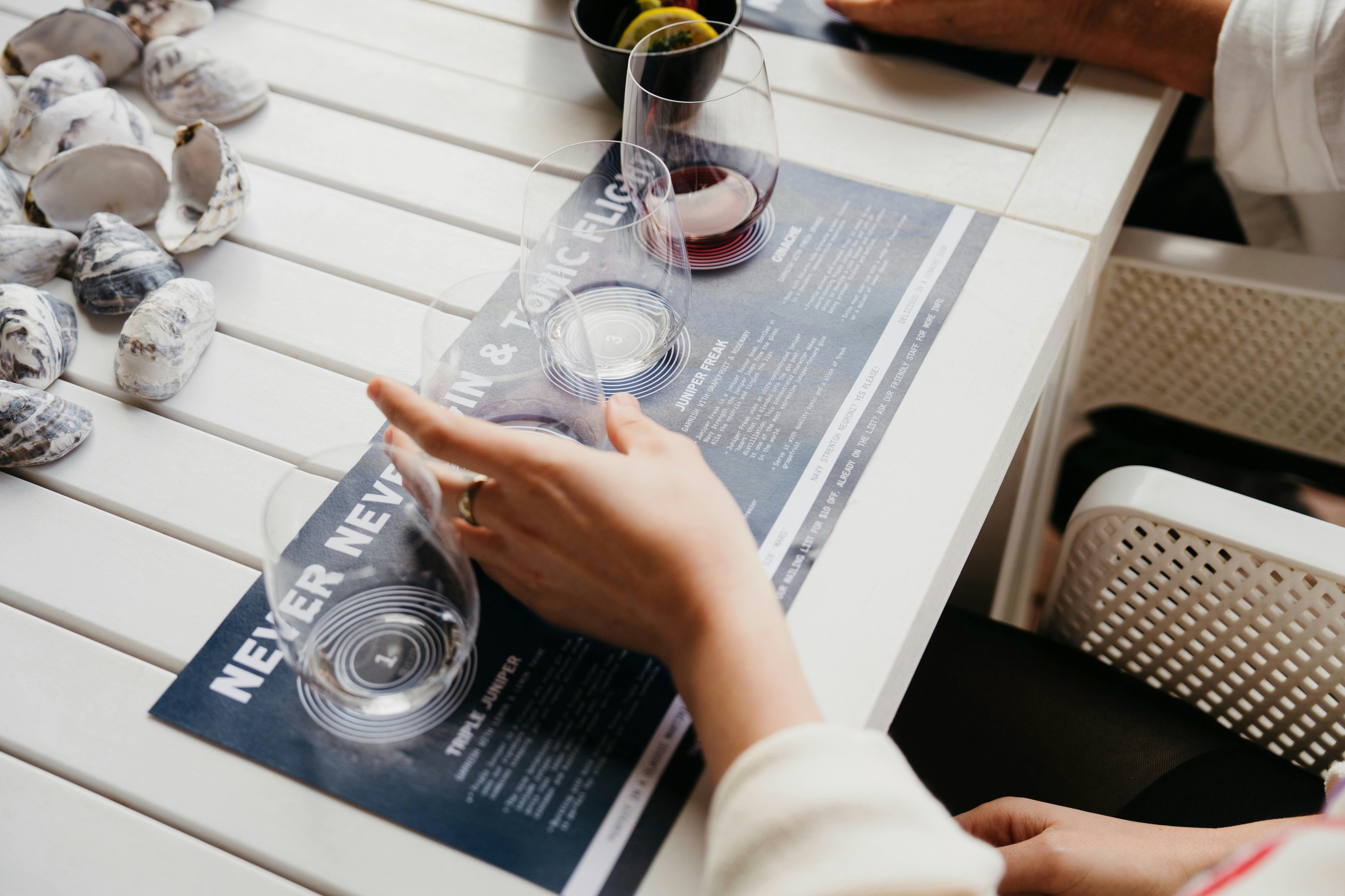 Hand holding tasting glass on gin flight mat with oyster shells on a white table.