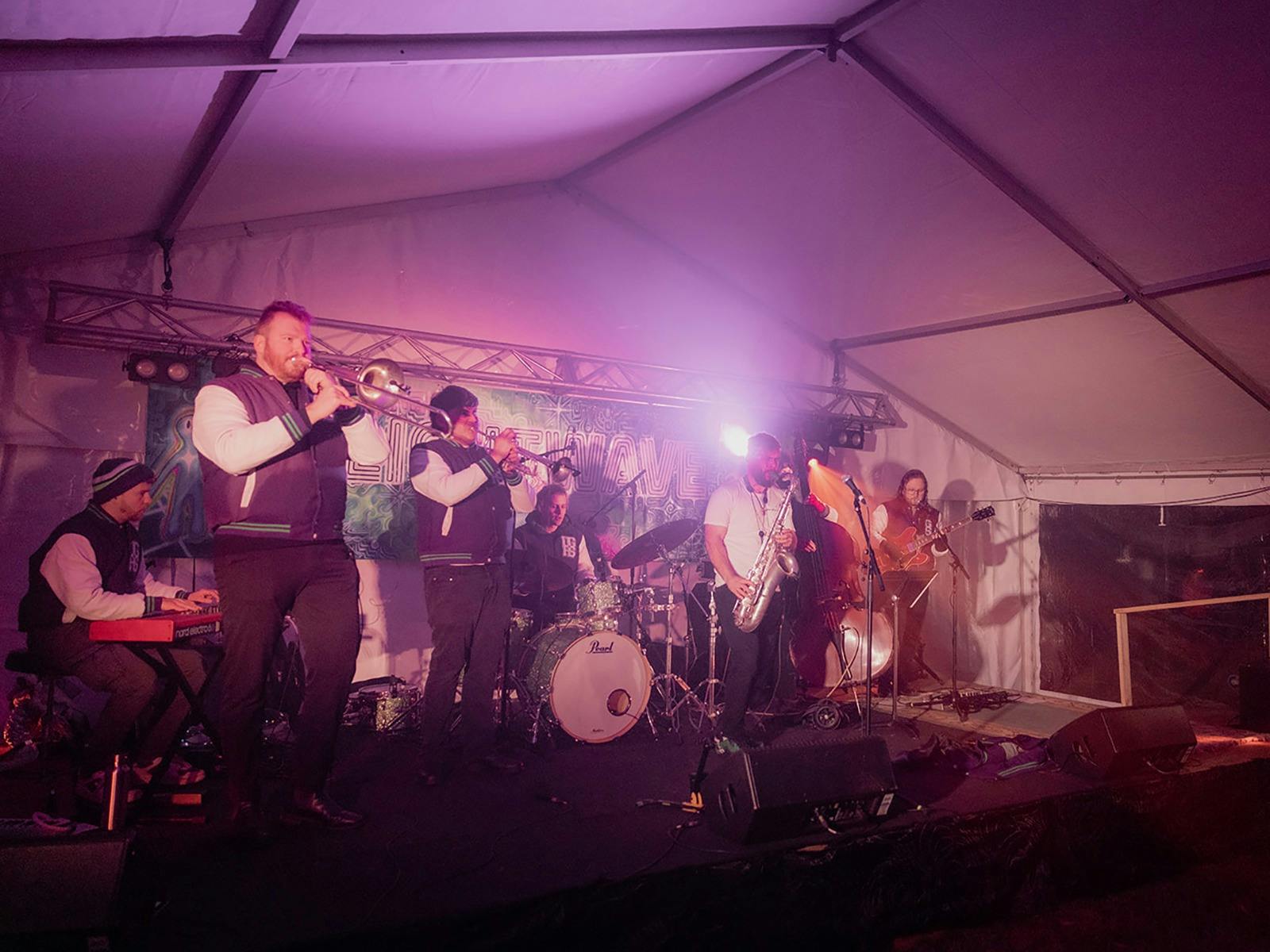 Band members in purple and white bomber jackets are playing  various instruments in a marquee.
