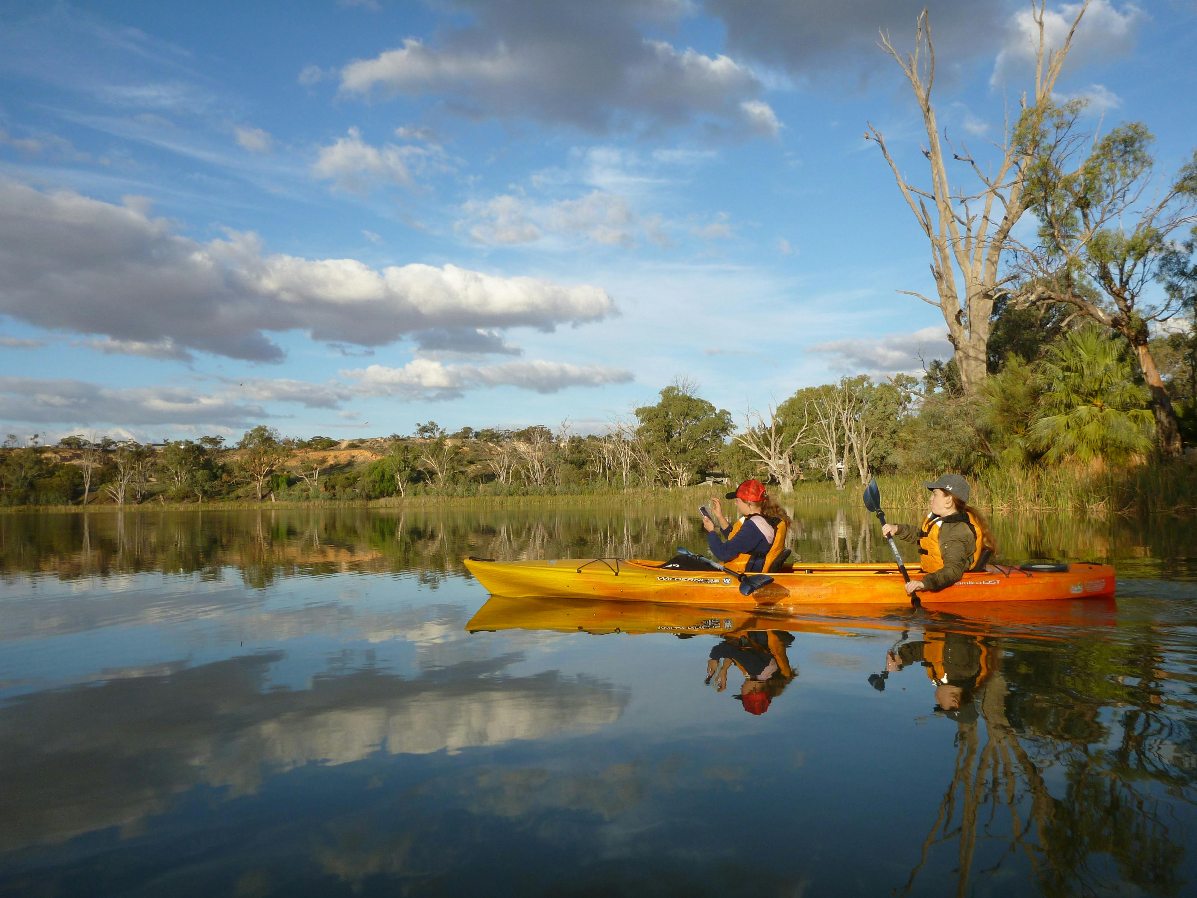 One kayaker photographs as another paddles a wetland lake with mirror-like reflections.