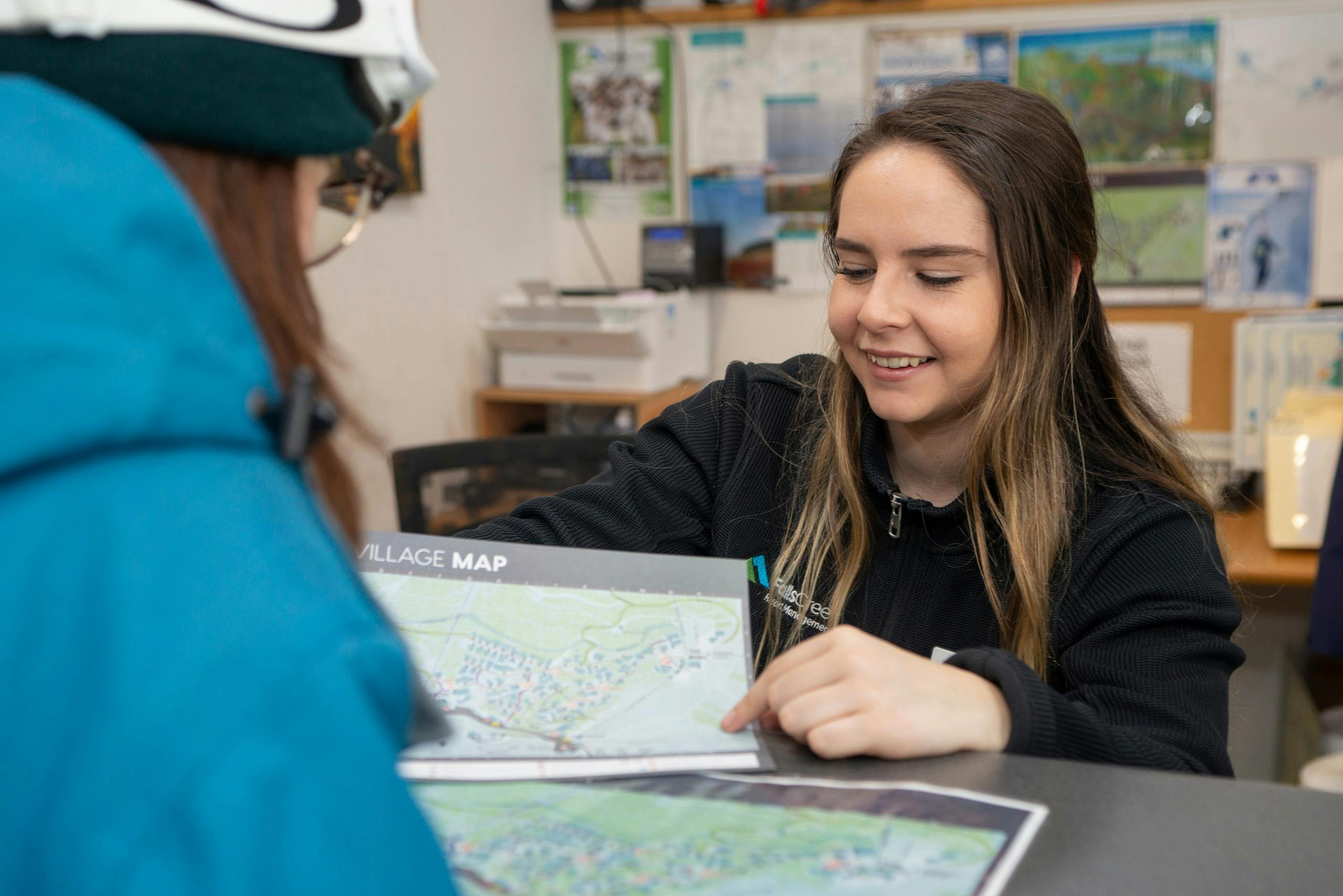Smiling young female staff member pointing to a Falls Creek village map, speaking to a guest.