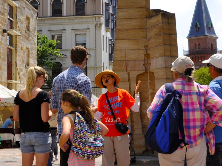 The Rocks free walking tour led by a tour guide in orange uniform is next to First Impressions art