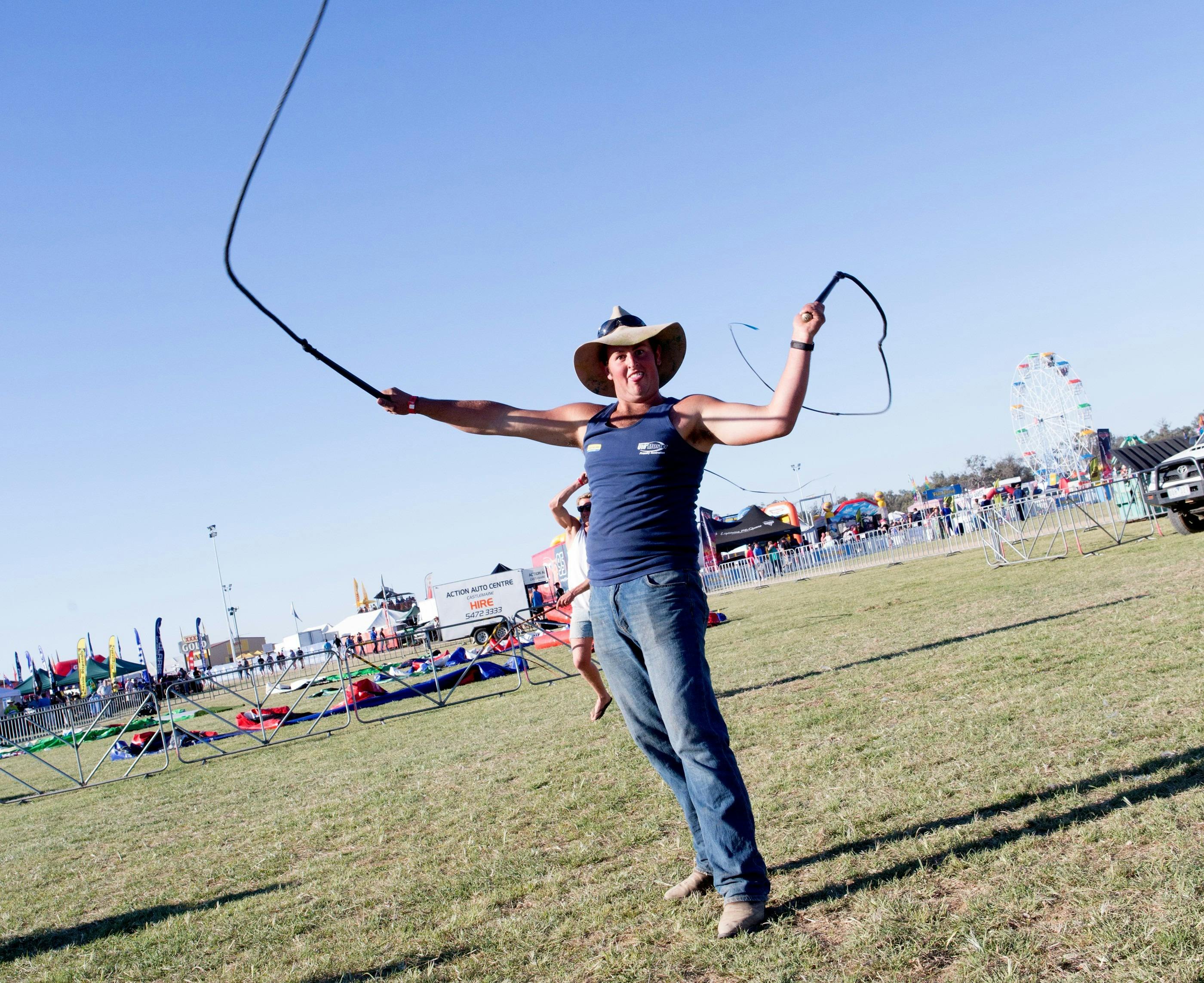 Man in Akubra hat, blue singlet and jeans cracks two whips on grass against a blue sky background