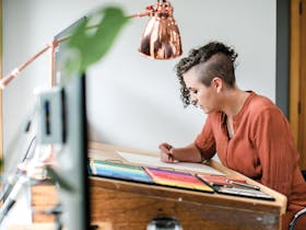 The artist sitting behind a desk, drawing. Coloured pencils on the desk, with a lamp and computer.