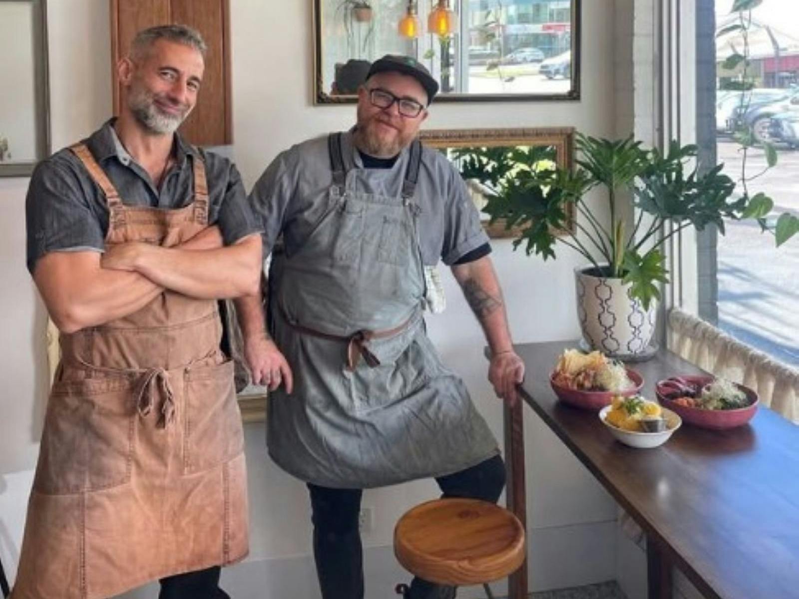 two Charcoal Bay owners standing in their aprons next to their food.