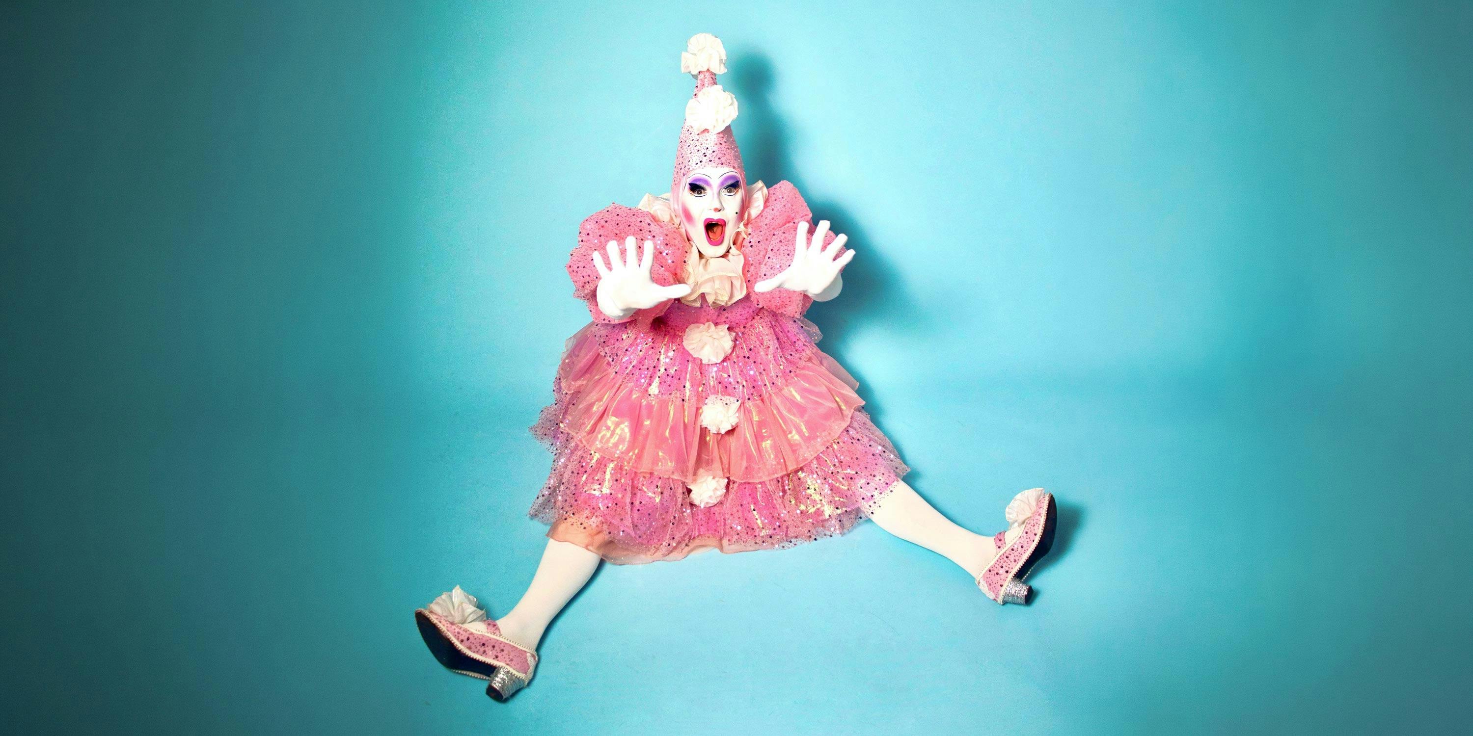 A drag performer in a pink clown outfit, sitting, holding their hands out to stop something