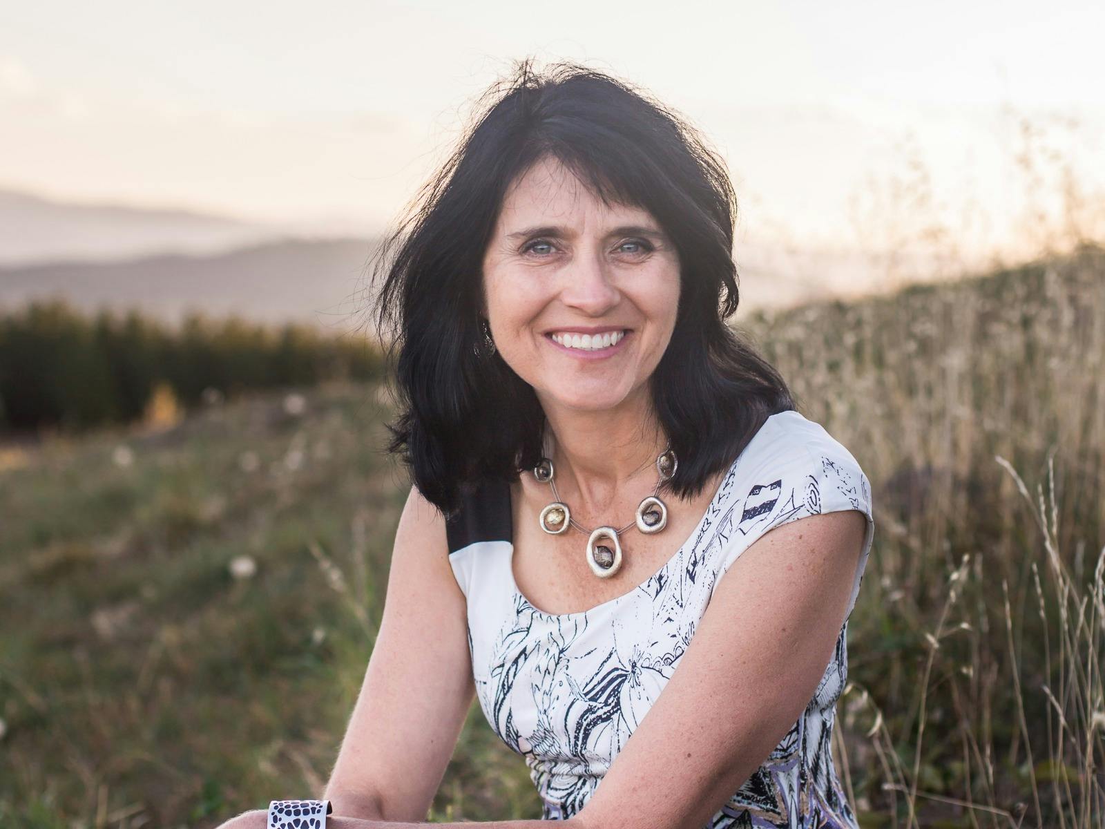 A woman with dark, wavy hair sitting outdoors, wearing a white patterned top.