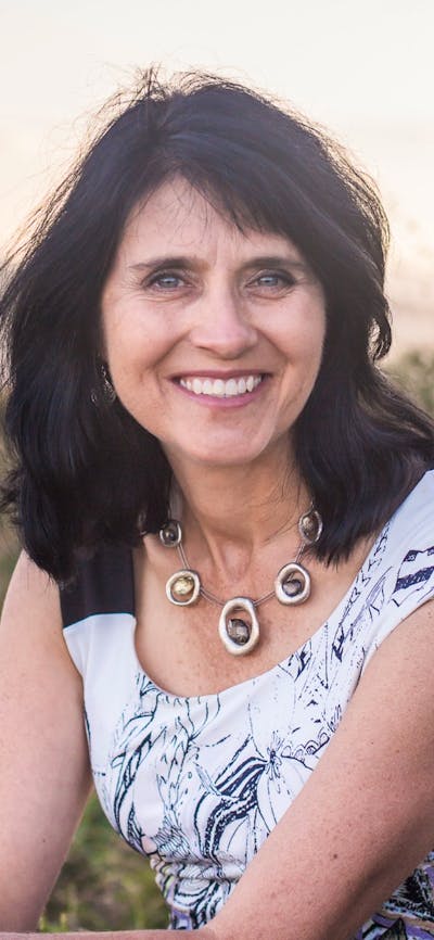 A woman with dark, wavy hair sitting outdoors, wearing a white patterned top.