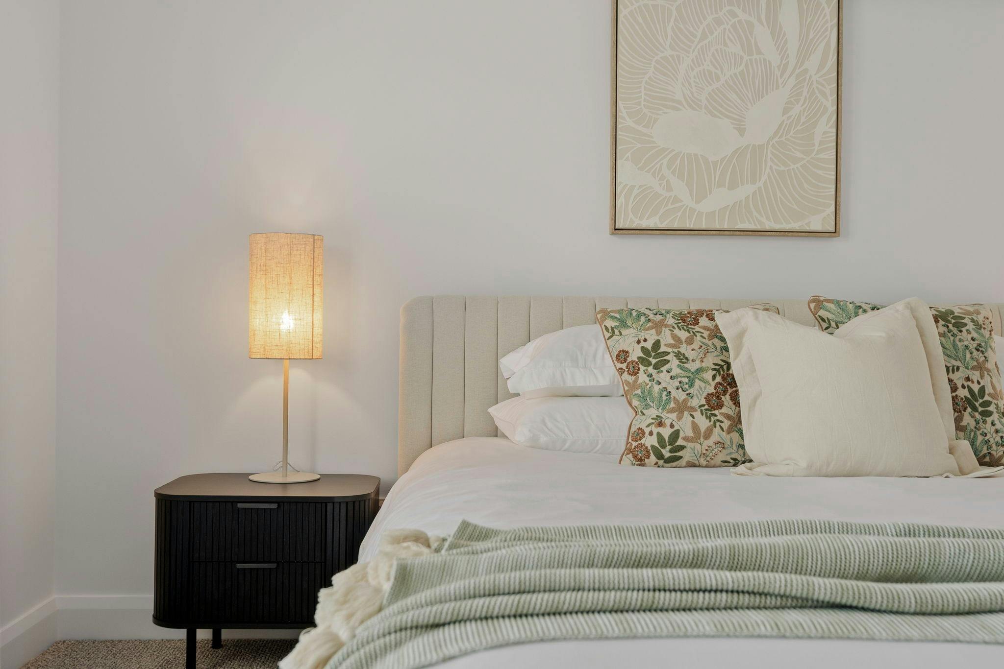 King bed with white linens, floral decor pillows, green throw, black side tables, lamps and art