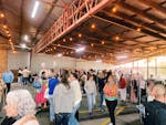 A lively indoor market with shoppers browsing clothes racks under warm, vibrant lighting.