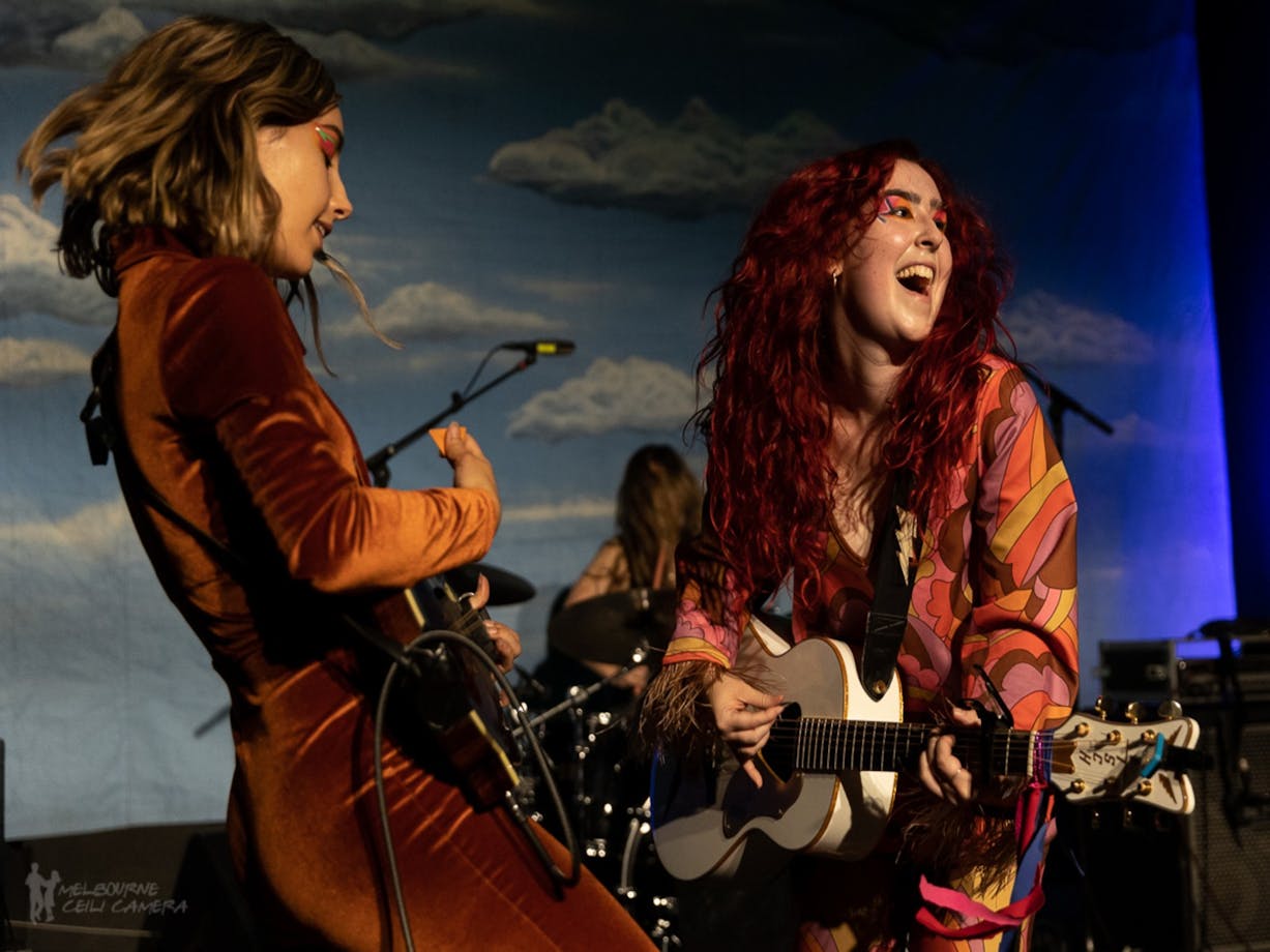 2 young woman dressed in fun outfits play guitars and sing against a sky blue backdrop
