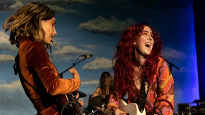 2 young woman dressed in fun outfits play guitars and sing against a sky blue backdrop