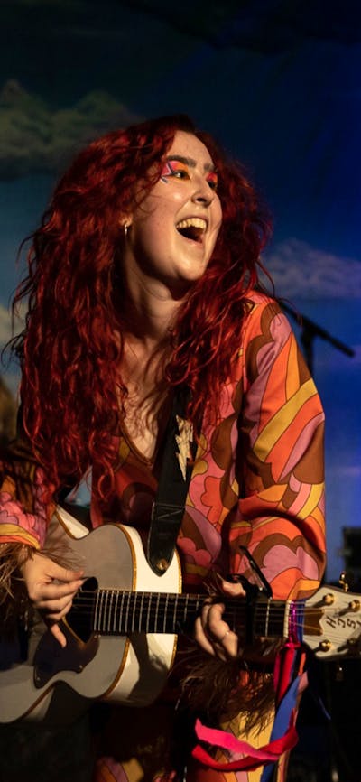 2 young woman dressed in fun outfits play guitars and sing against a sky blue backdrop