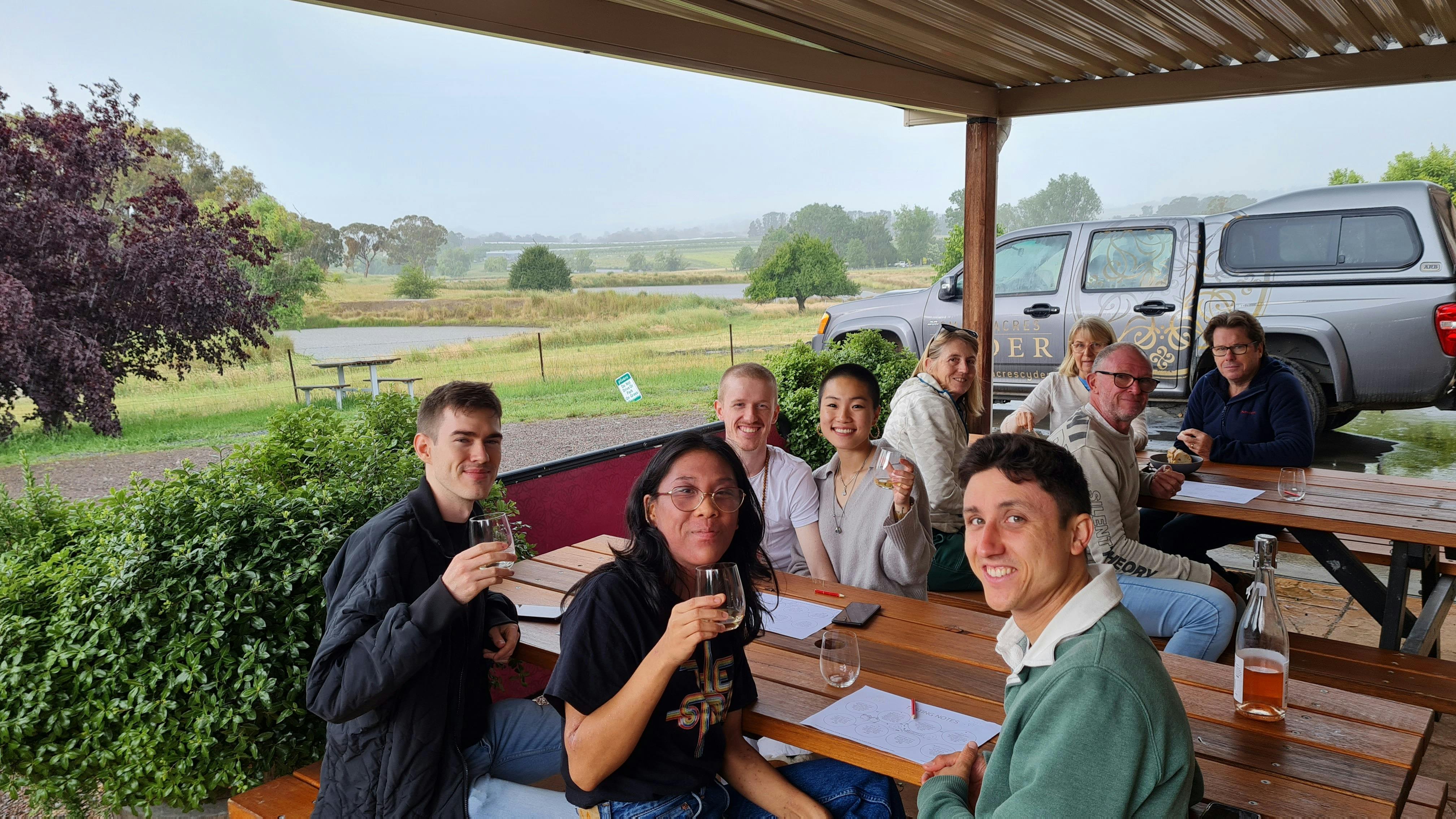 A group of people tasting cider on the veranda of Small Acres Cyder with views of the dam.