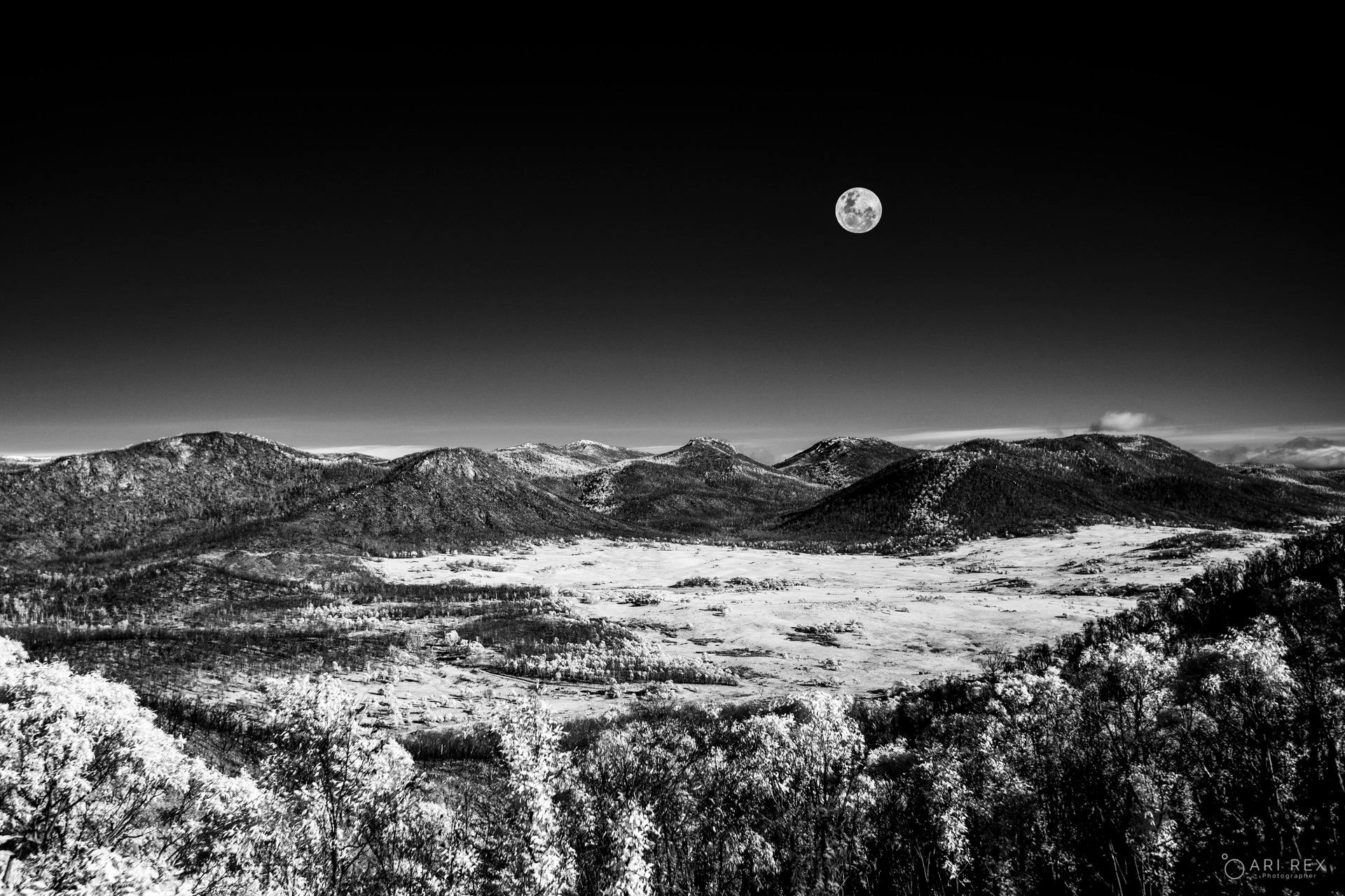 A small bright moon hangs above a wide, rugged mountain landscape in a black and white night photogr