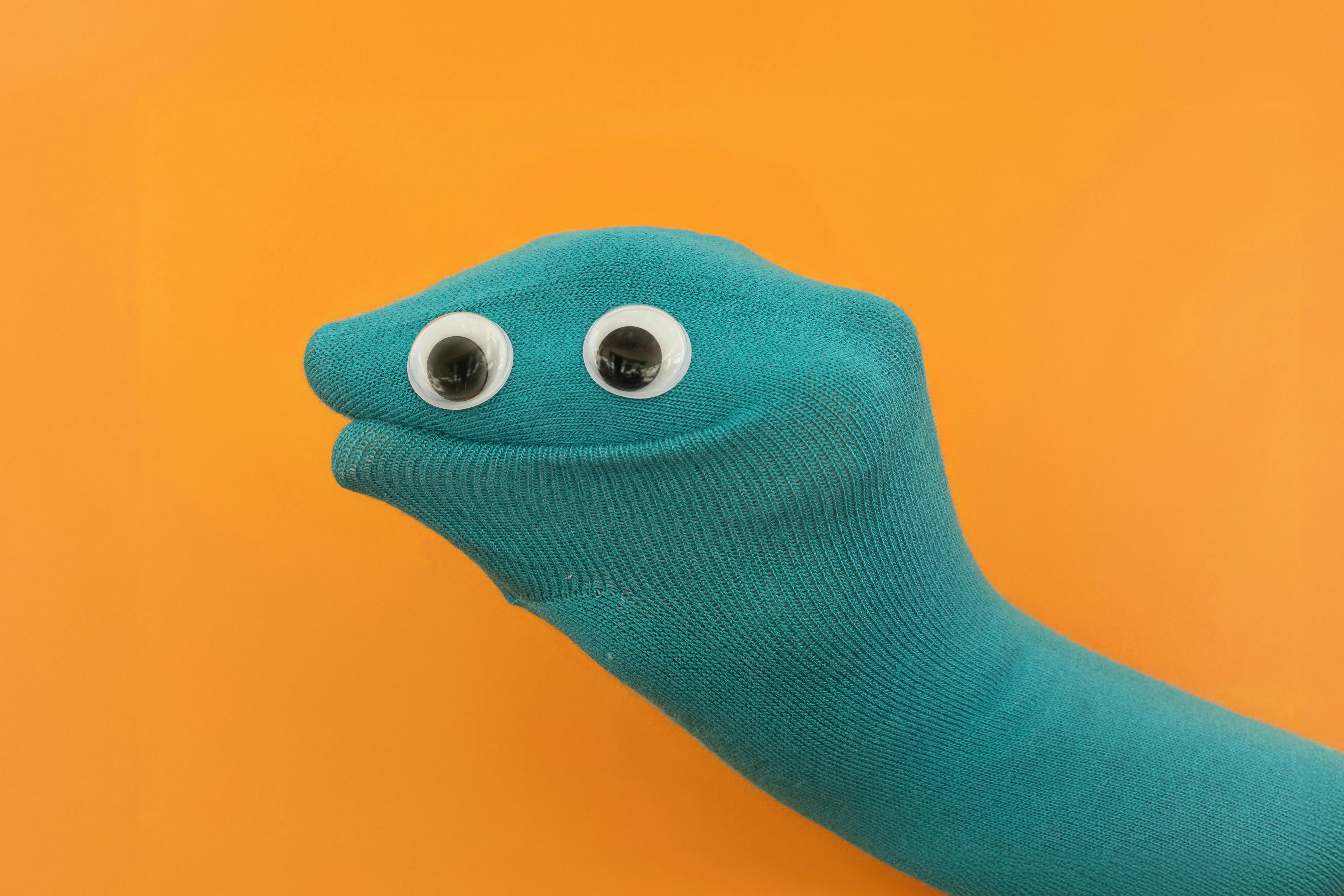A teal sock puppet with googly eyes sits against an orange background