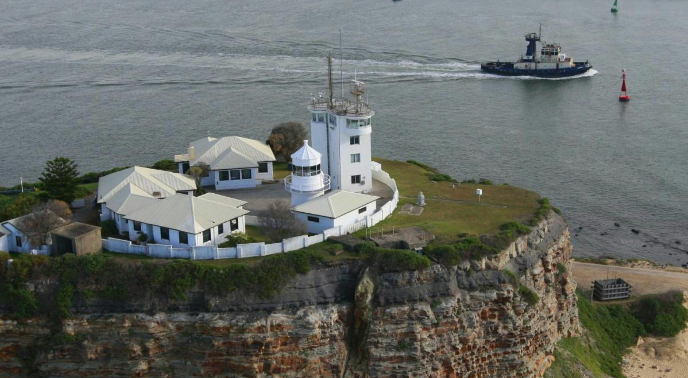 Nobbys Lighthouse close-up. You can see 3 cottages and the lighthouse. A tug boat is sailing past.