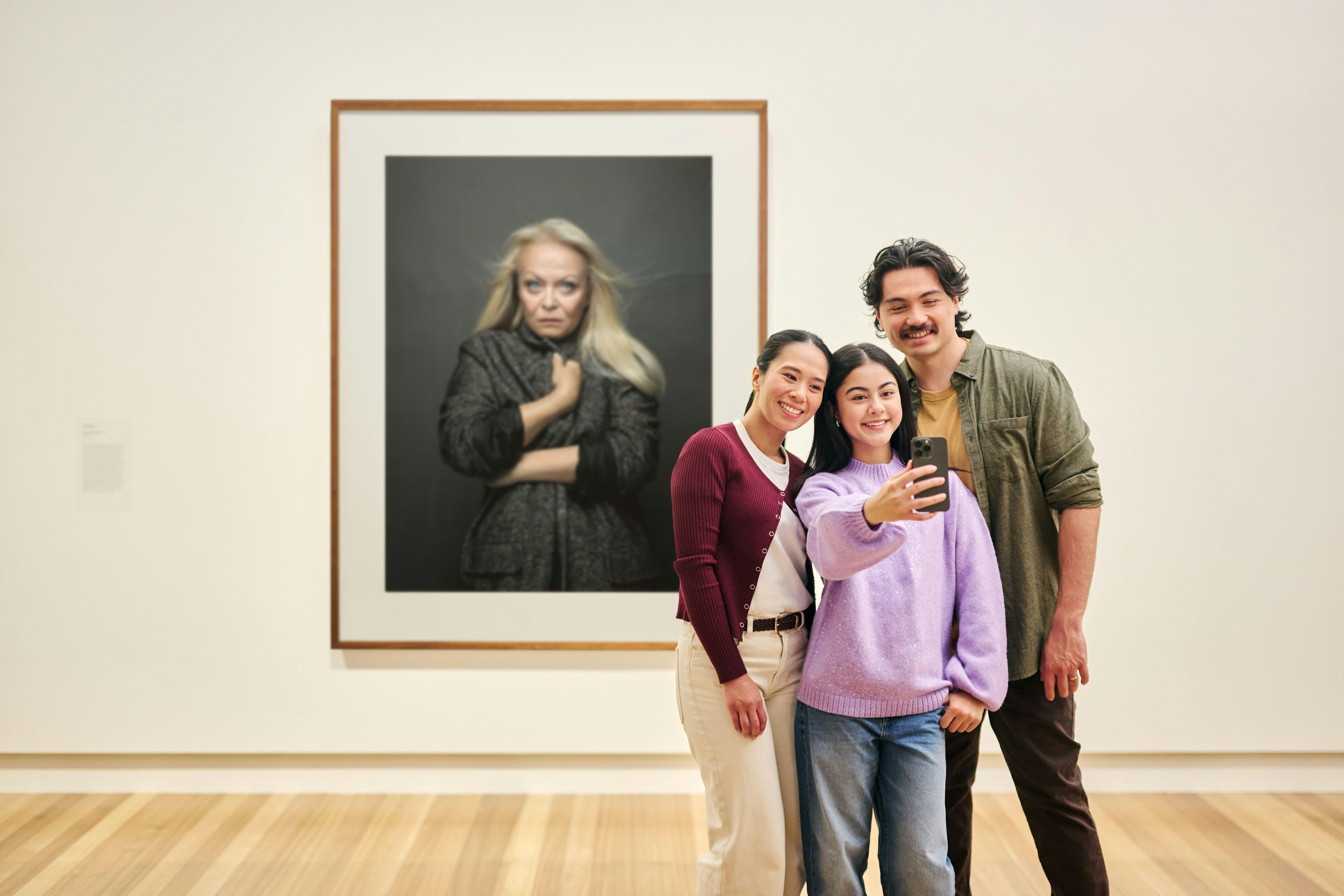 A family of three takes a selfie photo in front of a large portrait of actress Jacki Weaver.