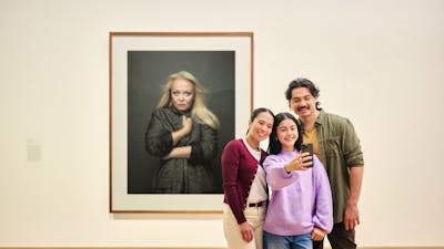 A family of three takes a selfie photo in front of a large portrait of actress Jacki Weaver.