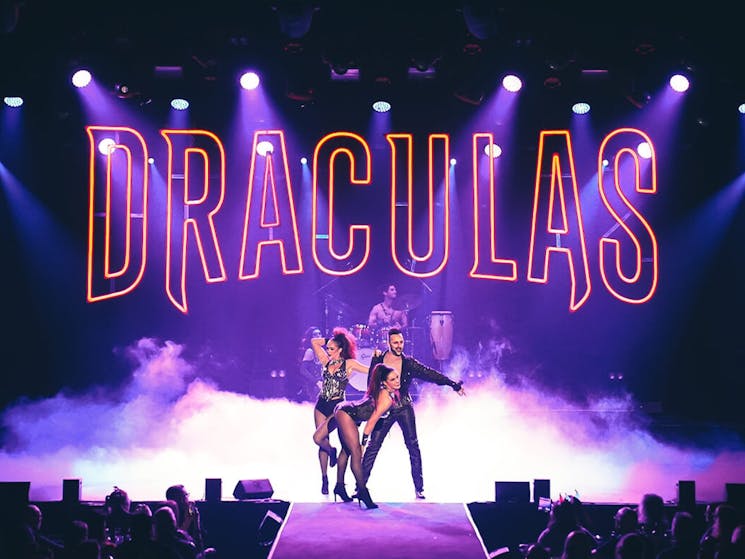 A male and two female performers dance under a large, red, neon Dracula's sign on stage.