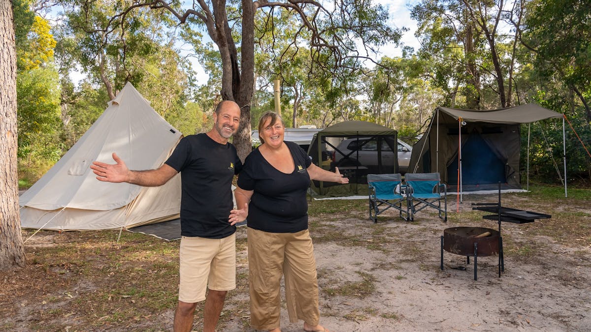 Two people standing with outstretched arms with setup camping tents behind them