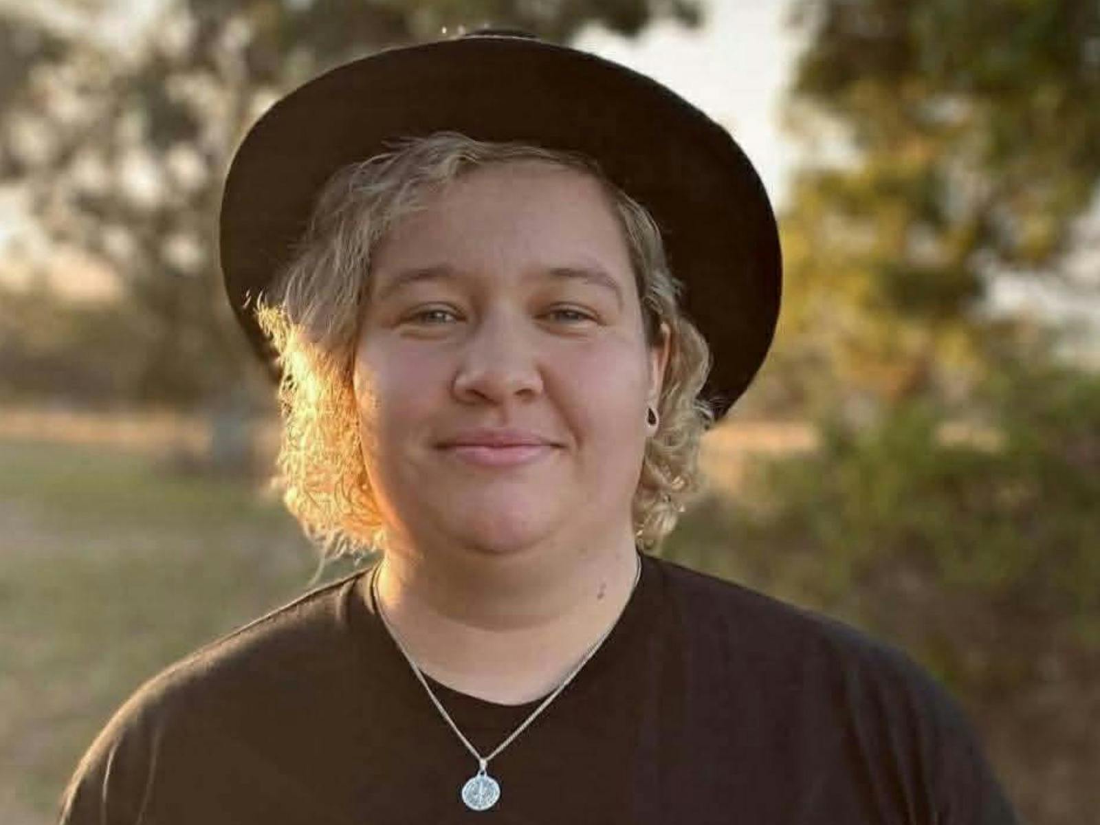 A person with curly blonde hair wearing a black hat and black t-shirt smiles at the camera.
