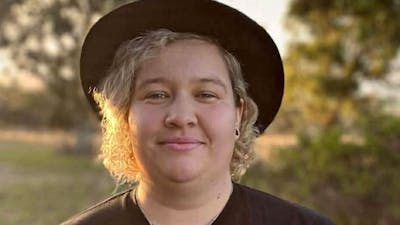 A person with curly blonde hair wearing a black hat and black t-shirt smiles at the camera.