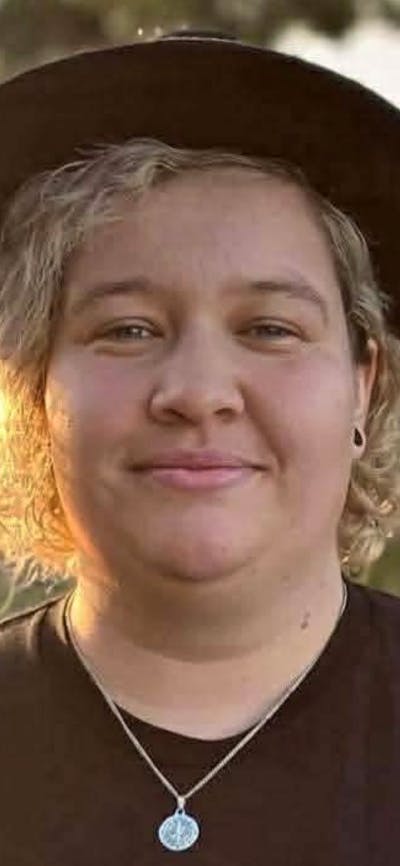 A person with curly blonde hair wearing a black hat and black t-shirt smiles at the camera.