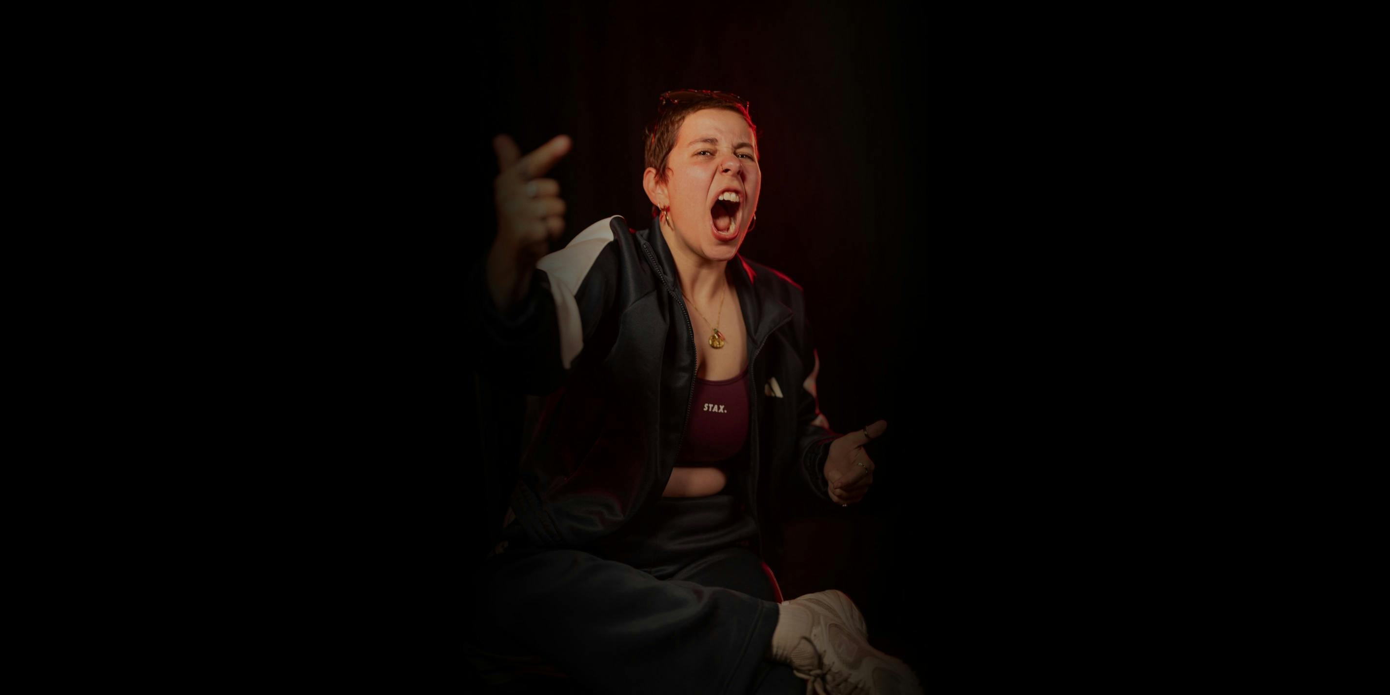 A performer mid-shout in a maroon top and black tracksuit jacket against a black background.