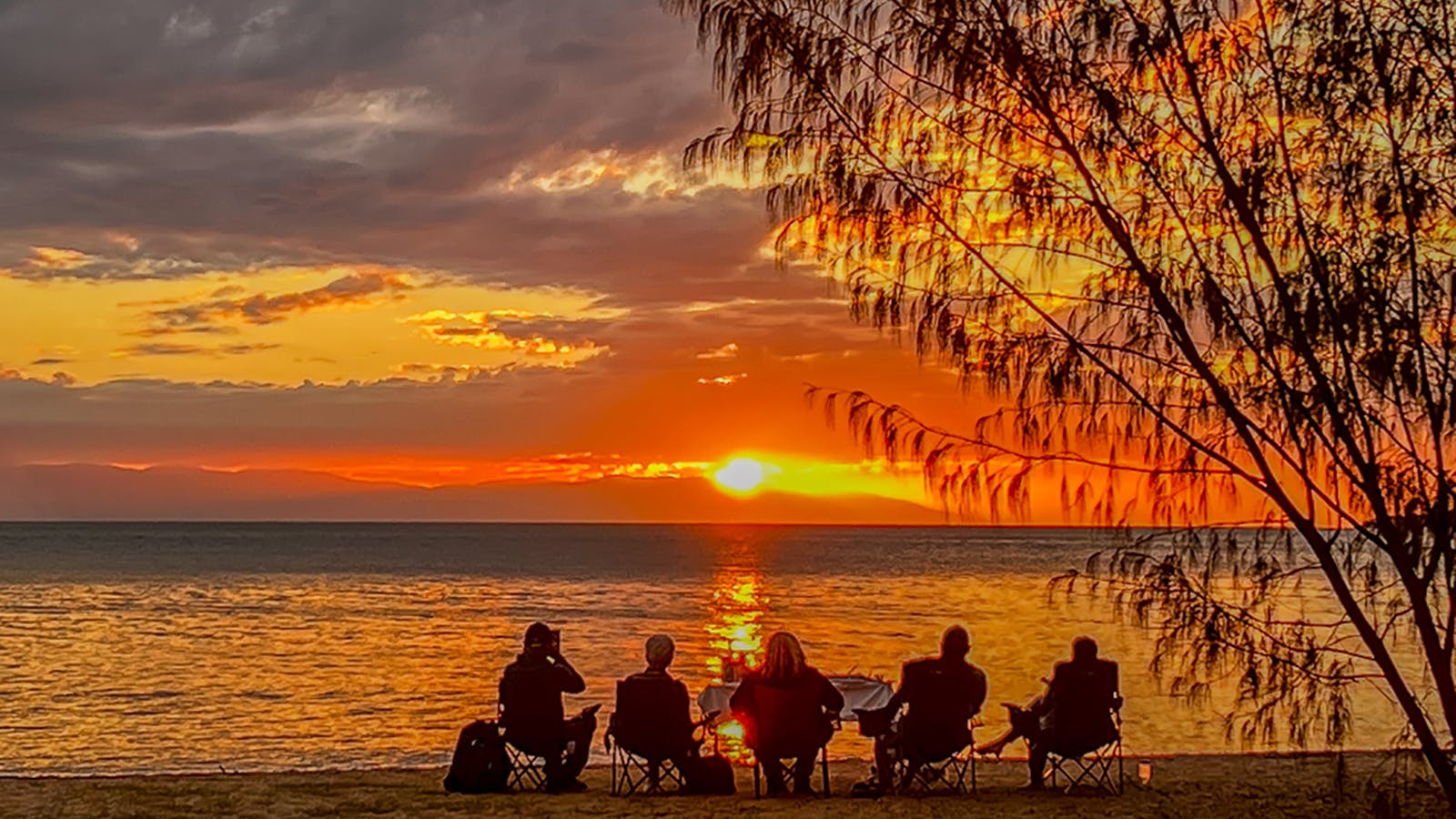 A group of five people sit on chairs at the beach watching a golden sunset over the water. A group of five people sit on chairs at the beach watching a golden sunset over the water.