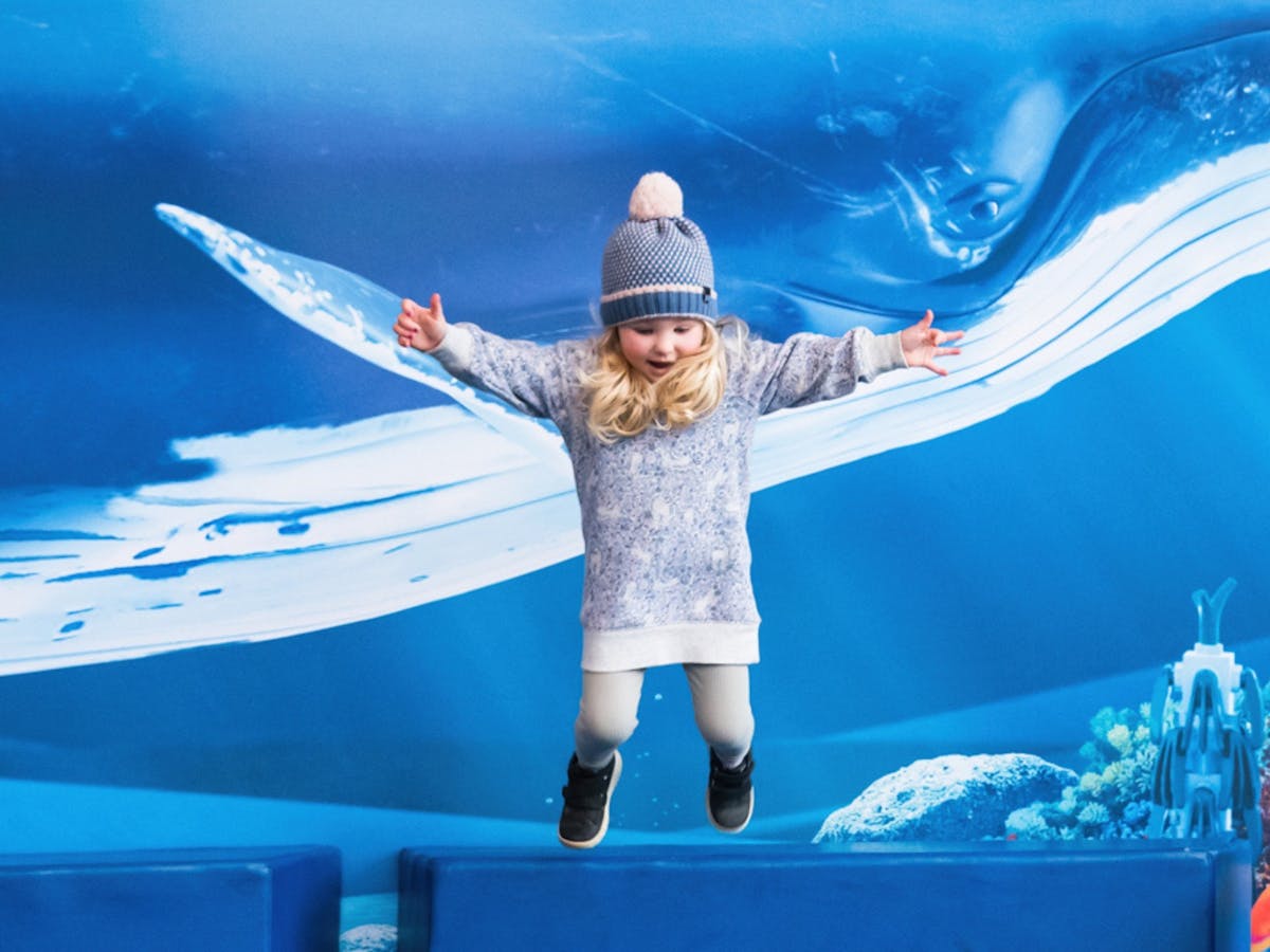 A young child in a beanie and jumper is jumping off a foam block with a picture of a whale behind.