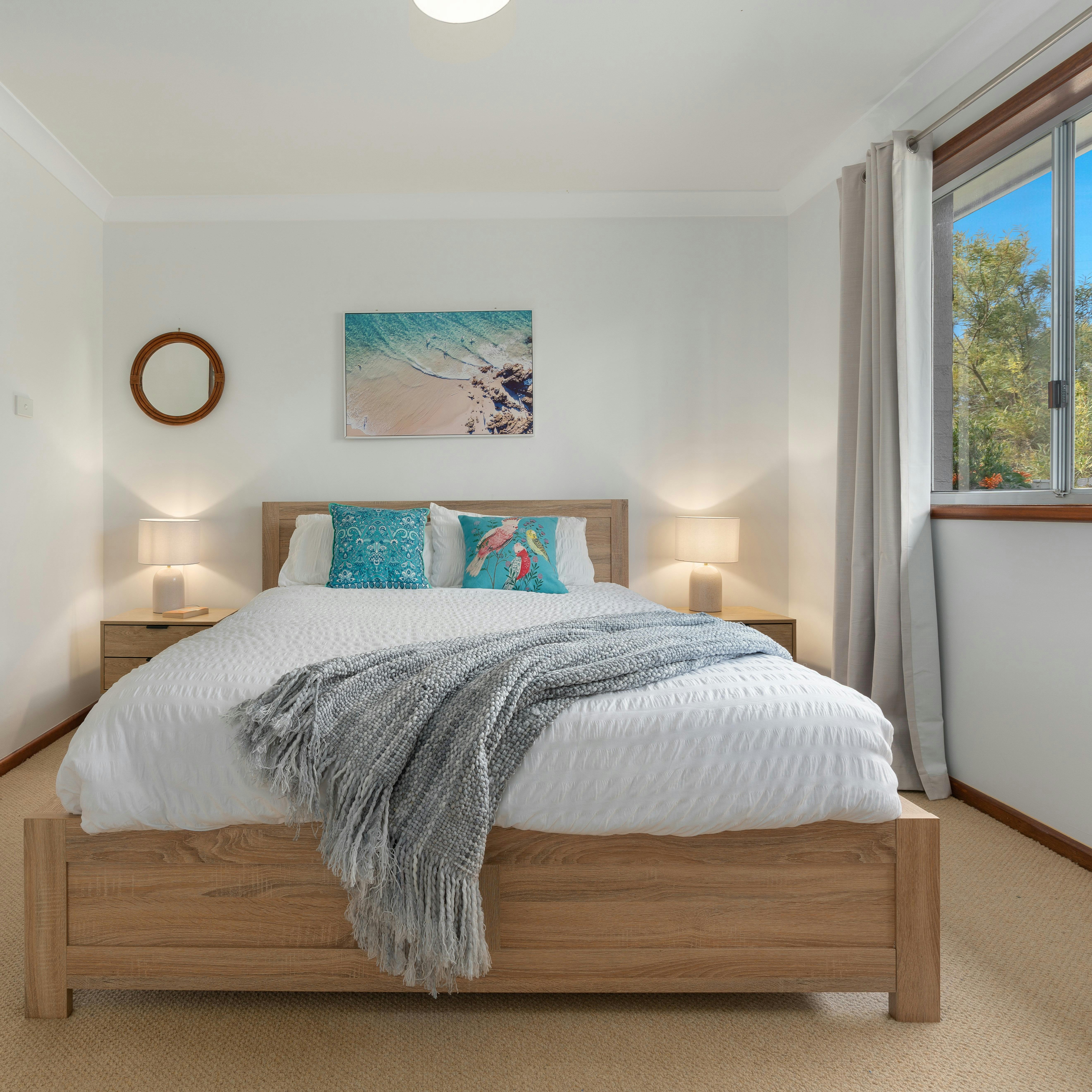 Queen Bed with white cover and blue cushions with bedside tables and lamps and coastal artwork on wa