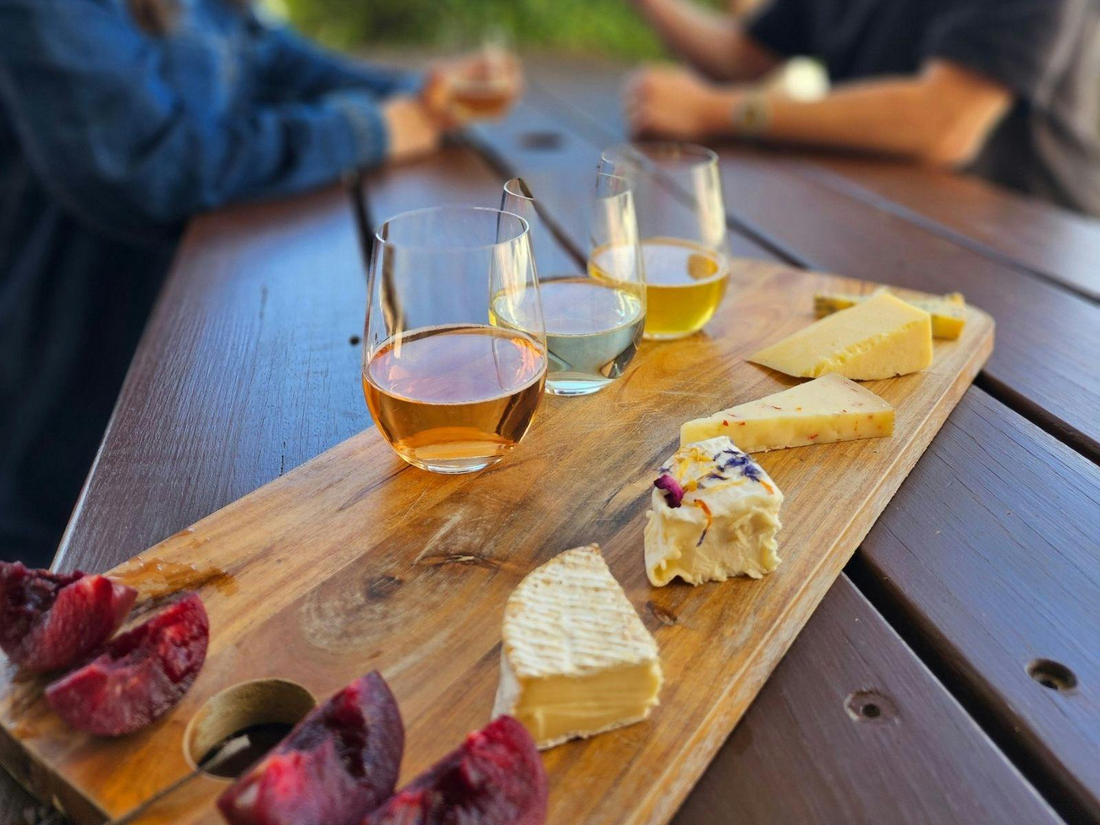 A wooden board with artisan cheeses and vintage ciders on a picnic table with a couple in background