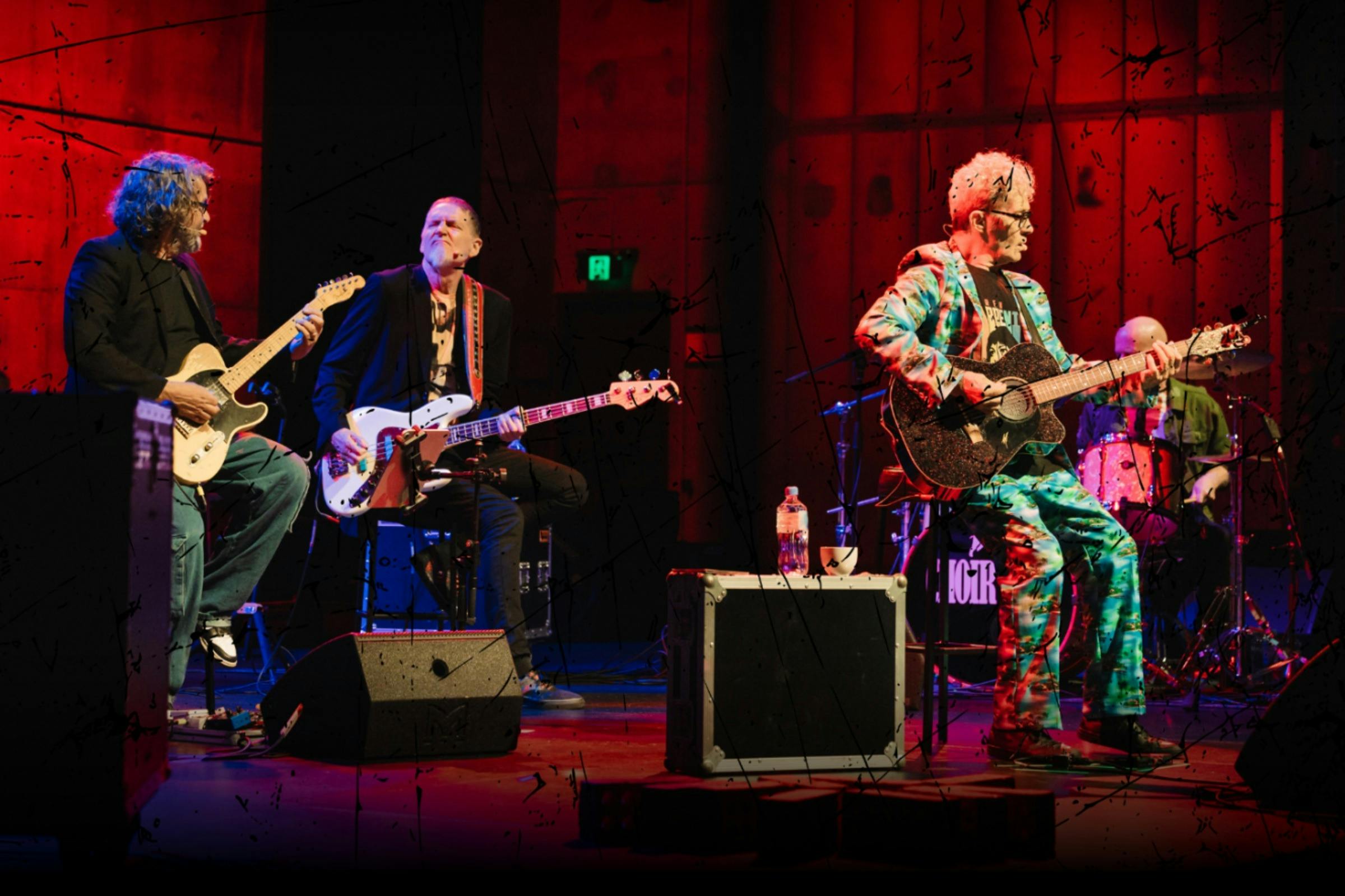 Four men on a stage, three are holding guitars and one is sat behind a drum kit
