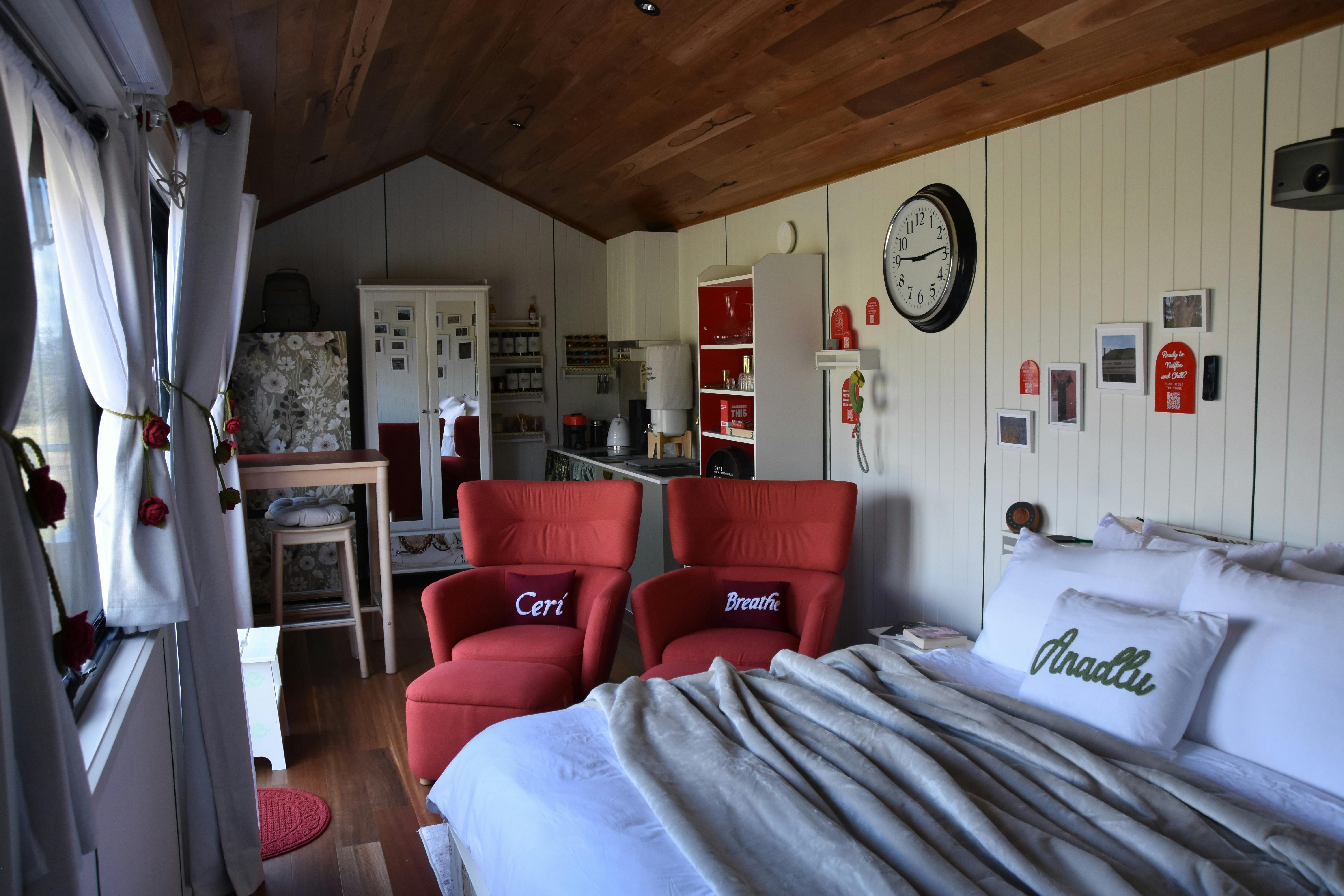 Interior of Ceri tiny home showing king bed, red armchairs, timber ceiling and styled decor