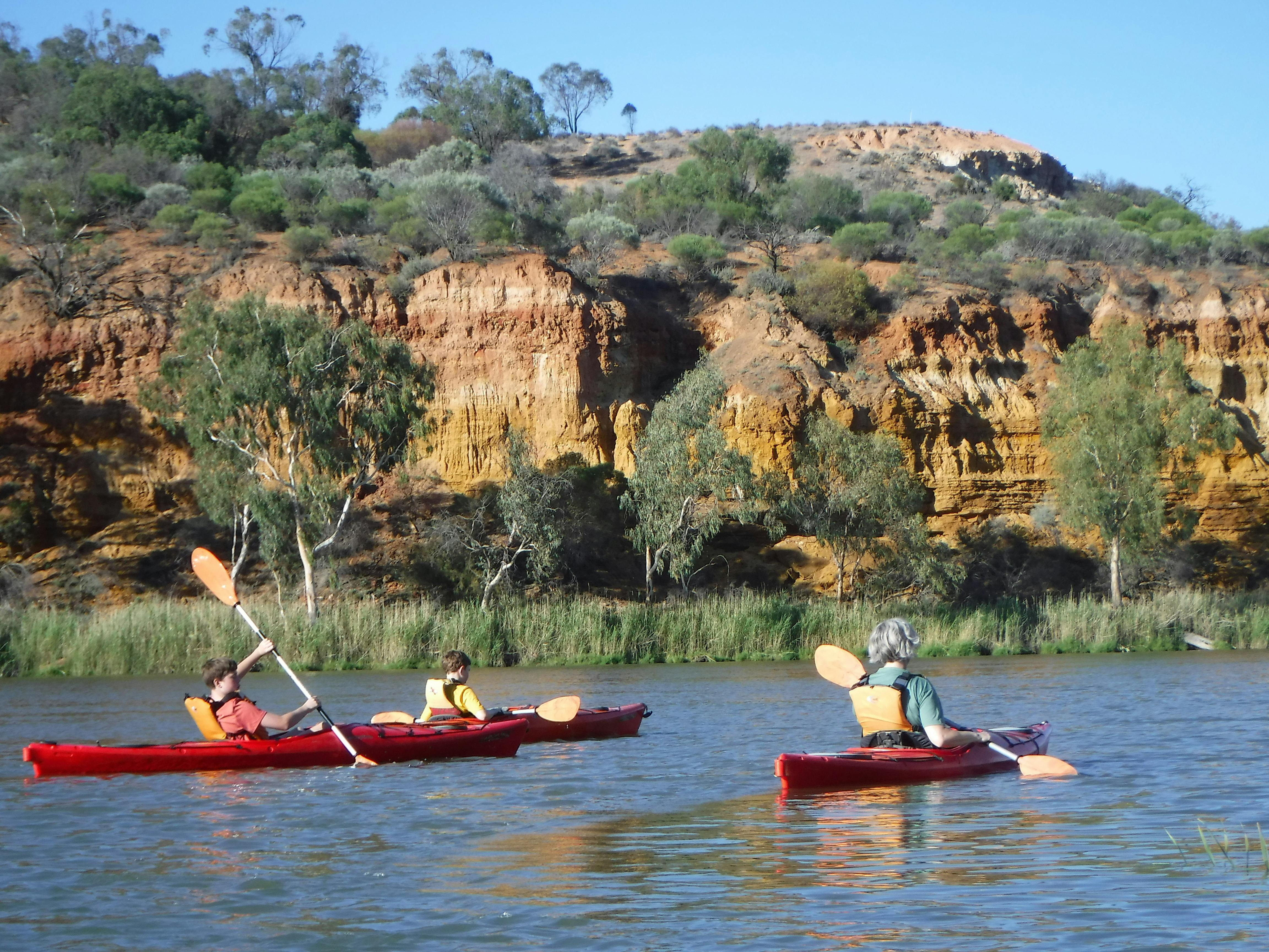 3 Kayakers paddle past colourful striped limestone cliffs.