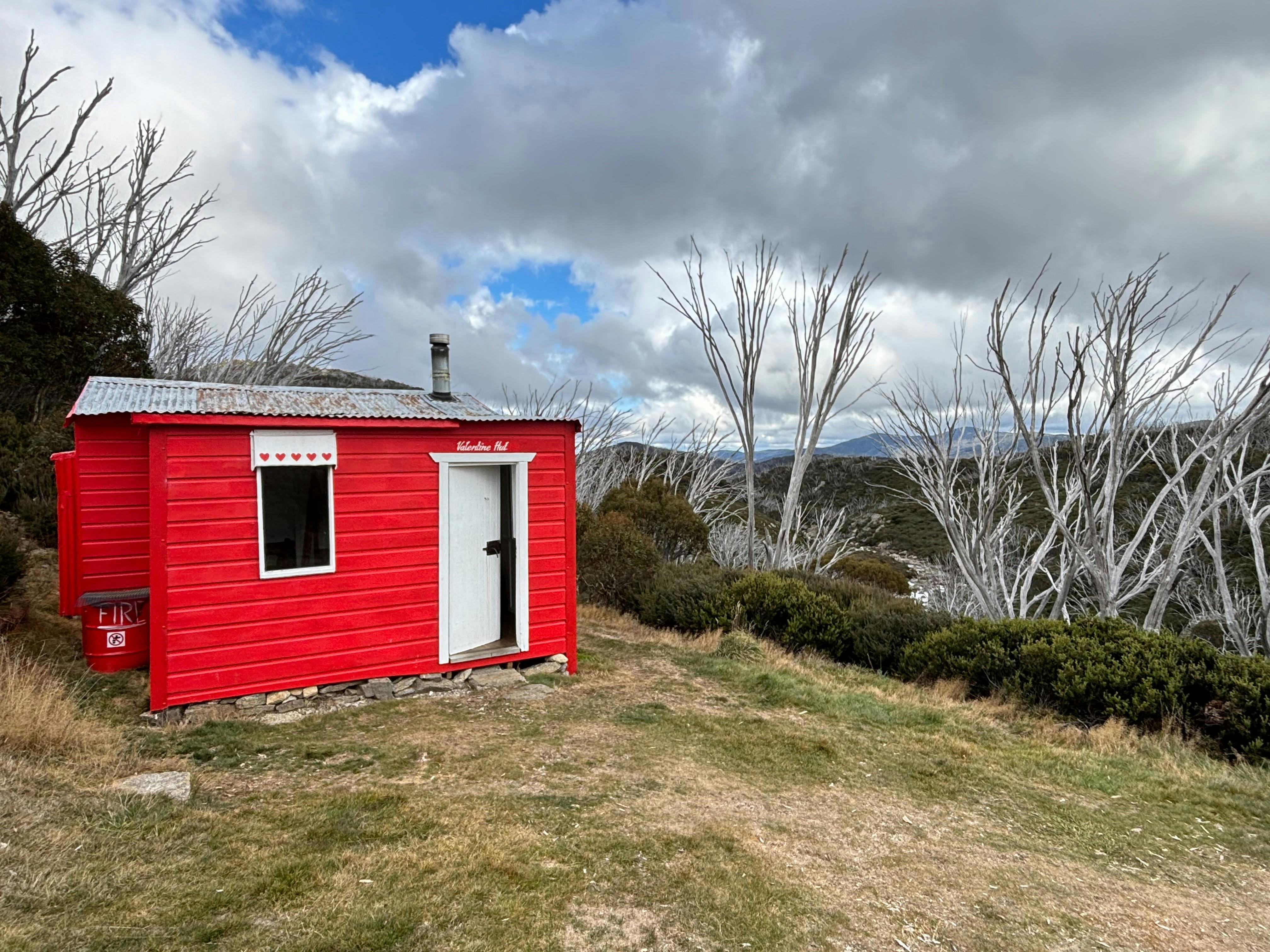 A small bright red hut sitting amongst snow gums on the side of Valentine River.