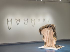 In an exhibition, several necklaces hang on the wall and a fur coat is draped in the foreground.