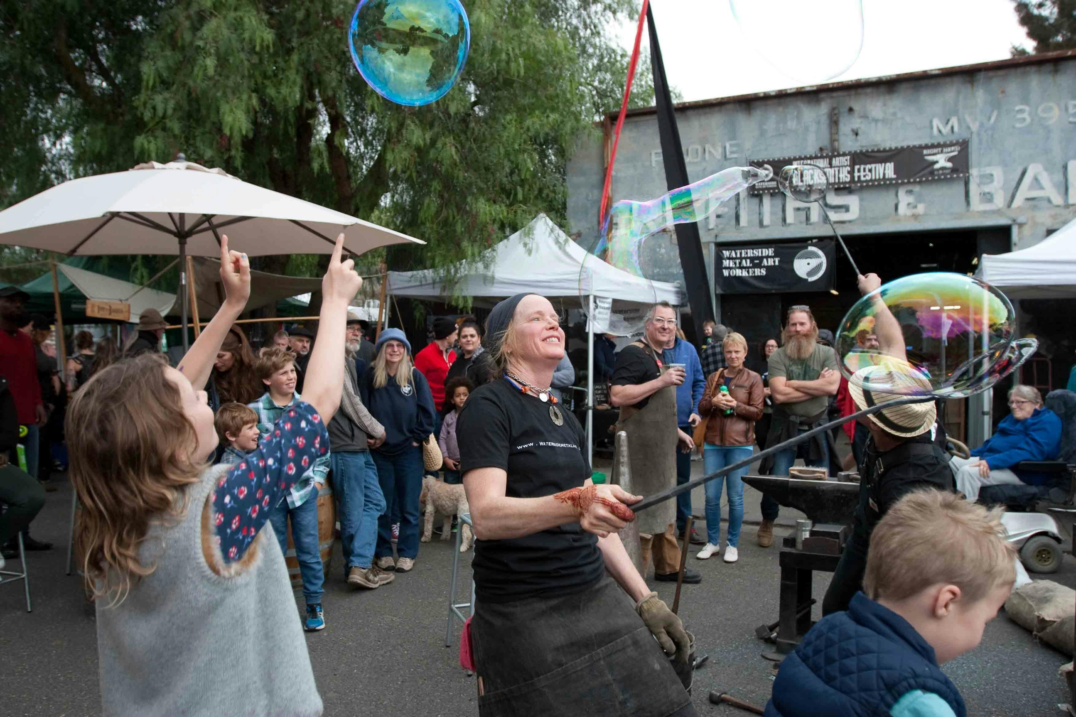 A blacksmith blows bubbles with a hand-forged wand as excited kids jump and reach for them.