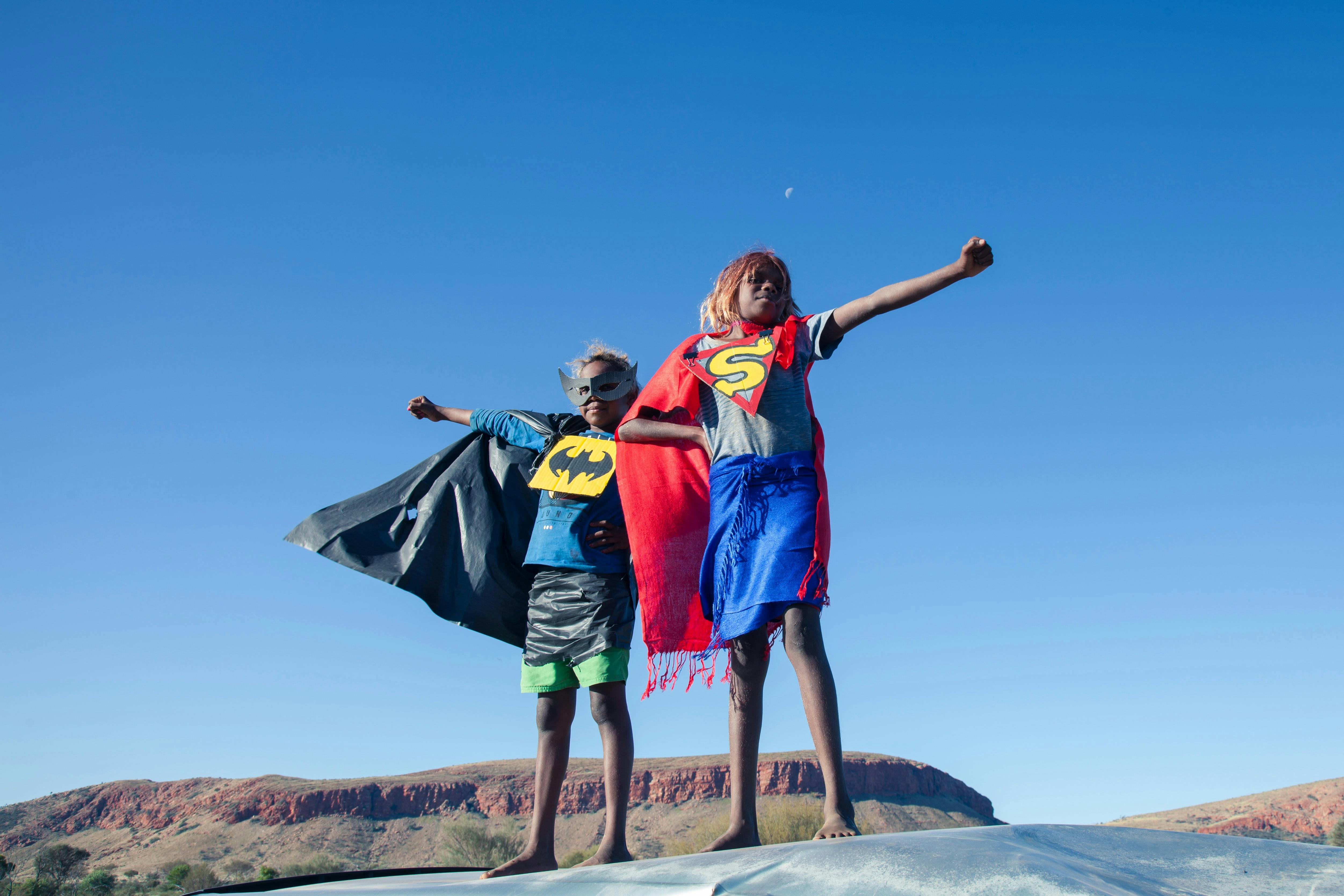 Photograph of two children standing on a rock under clear blue sky, dressed in superhero costumes.