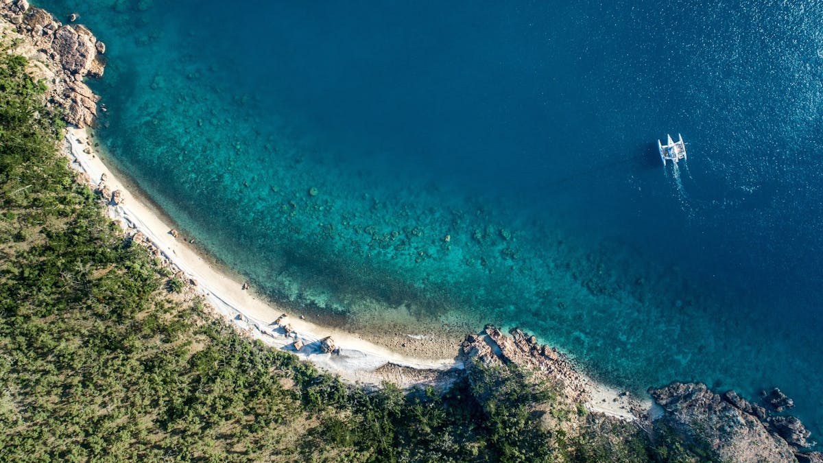 Aerial view of Blue Pearl Bay with turquoise waters, white sand, lush greenery, and a boat.